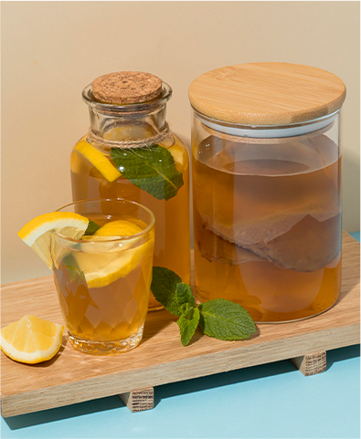Verre de thé glacé avec une tranche de citron, carafe en verre avec bouchon en liège contenant des feuilles de menthe et des tranches de citron, et pot en verre avec un couvercle en bois sur une planche en bois clair.