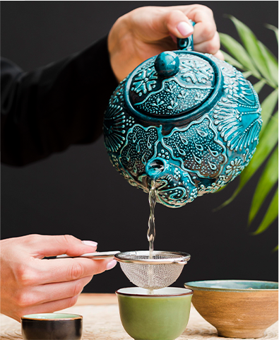 Une main verse du thé d’une théière bleue décorée dans une passoire tenue au-dessus d’une tasse verte.