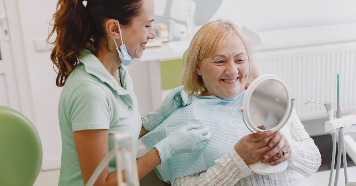 Older woman in a dental chair smiling into a hand mirror beside her dentist