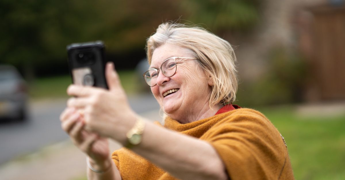 Smiling woman with glasses taking a photo of herself with a phone outdoors