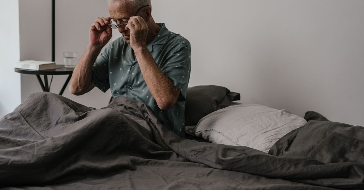 Man sitting up in bed in the morning rubbing his eyes under his glasses