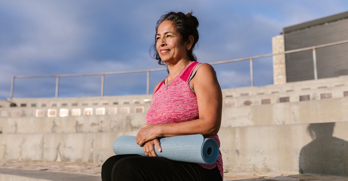 Woman in her fifties smiling outdoors in the city holding a yoga mat
