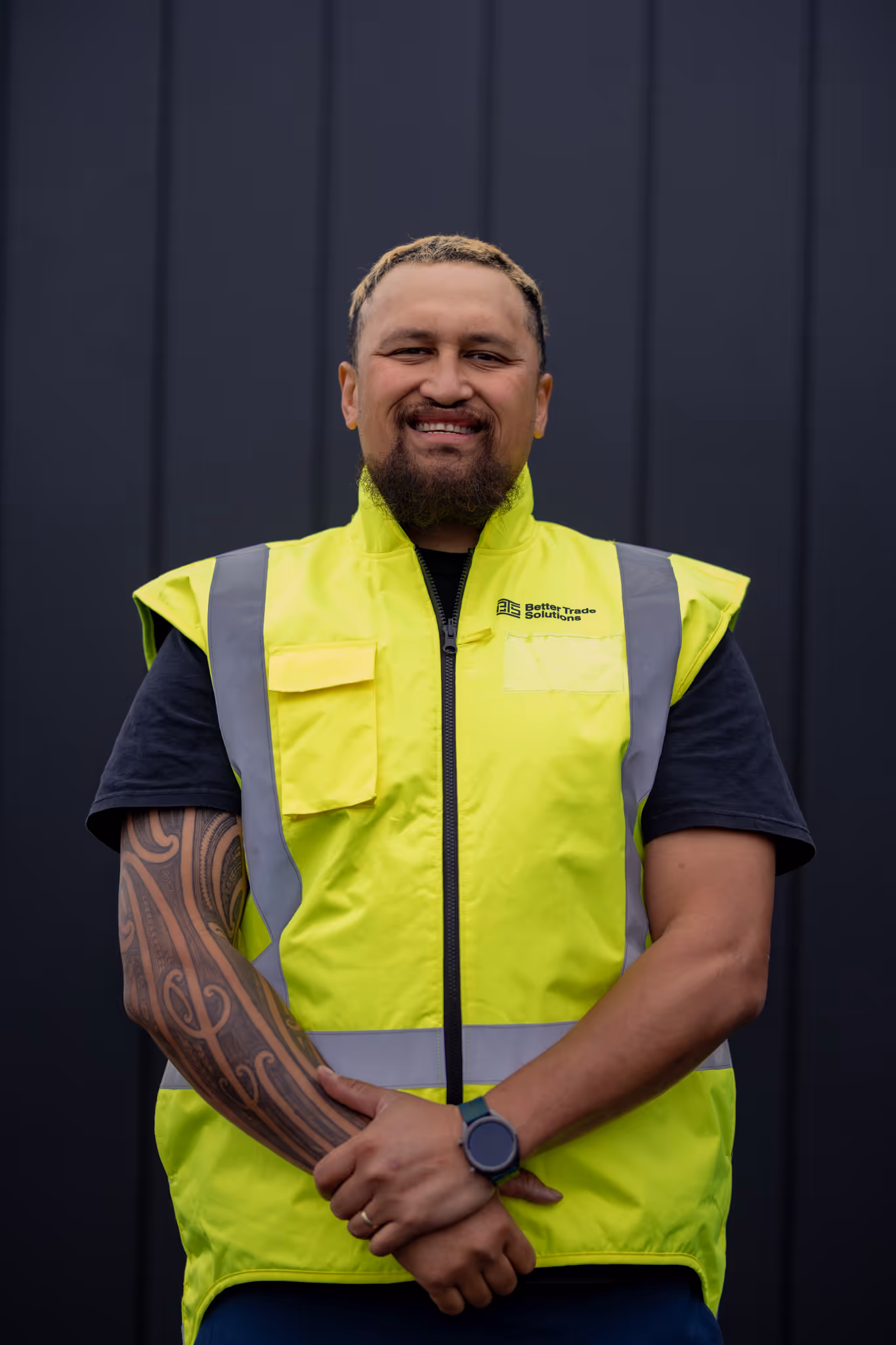 Smiling man wearing a yellow high-visibility vest with Better Trade Solutions logo and a watch, standing against a dark background.