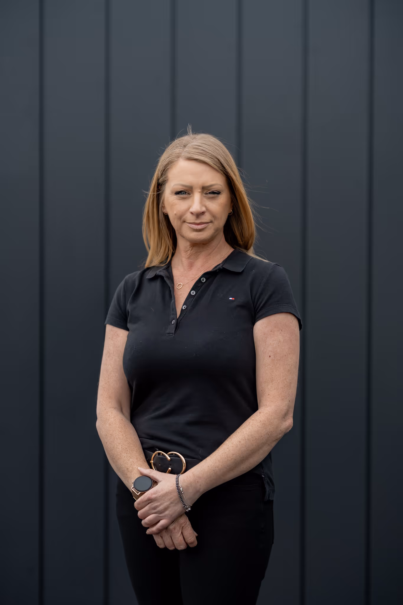 Woman with straight blonde hair in a black polo shirt and black pants standing in front of a dark panelled wall.