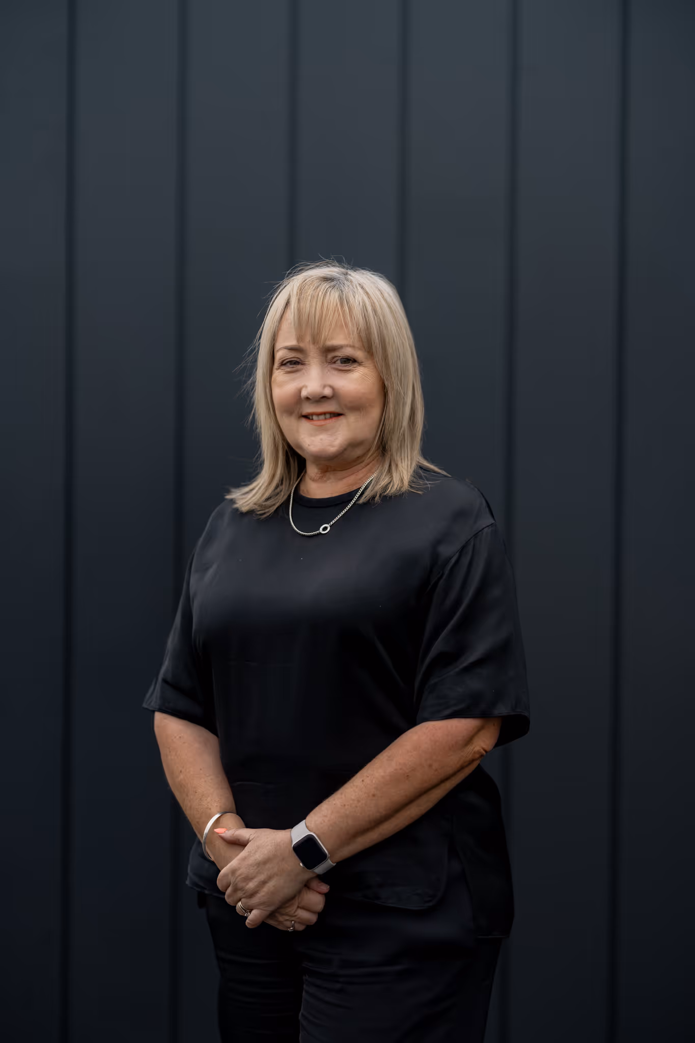 Smiling middle-aged woman with blonde hair wearing a black shirt and smartwatch standing against a dark paneled wall.