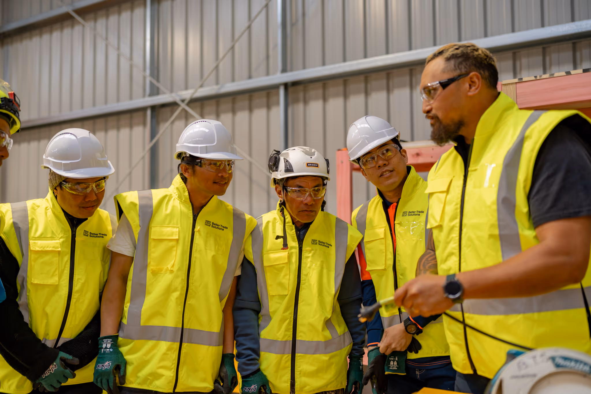 Construction workers in yellow high-visibility vests and helmets listening to a man explaining with a tool in a warehouse.