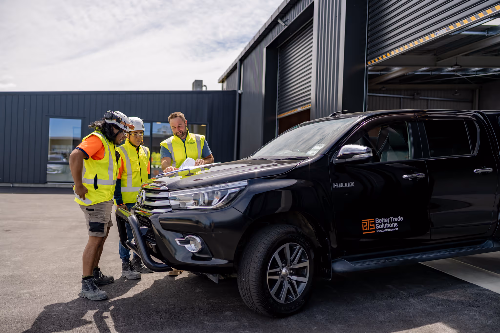Three construction workers wearing high-vis vests and helmets review a document beside a black Hilux ute branded with Better Trade Solutions outside a warehouse.