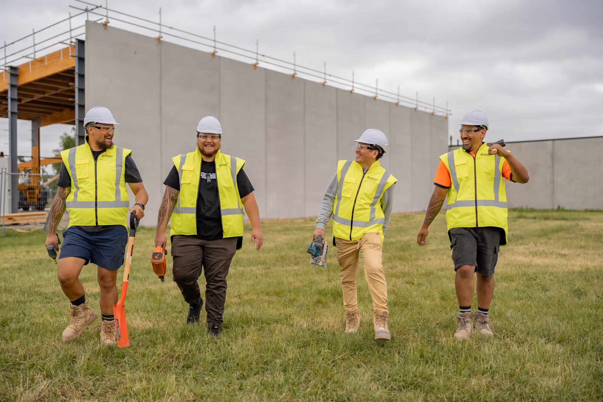 Four construction workers wearing white helmets and yellow safety vests walking on grass near a building under construction, smiling and carrying tools.