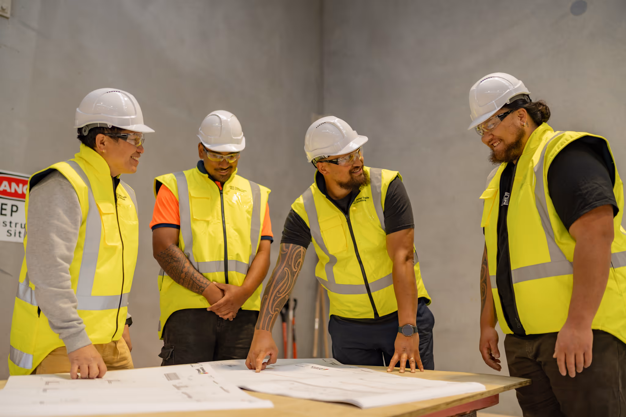 Four construction workers in safety helmets and yellow vests reviewing blueprints on a table inside a building site.