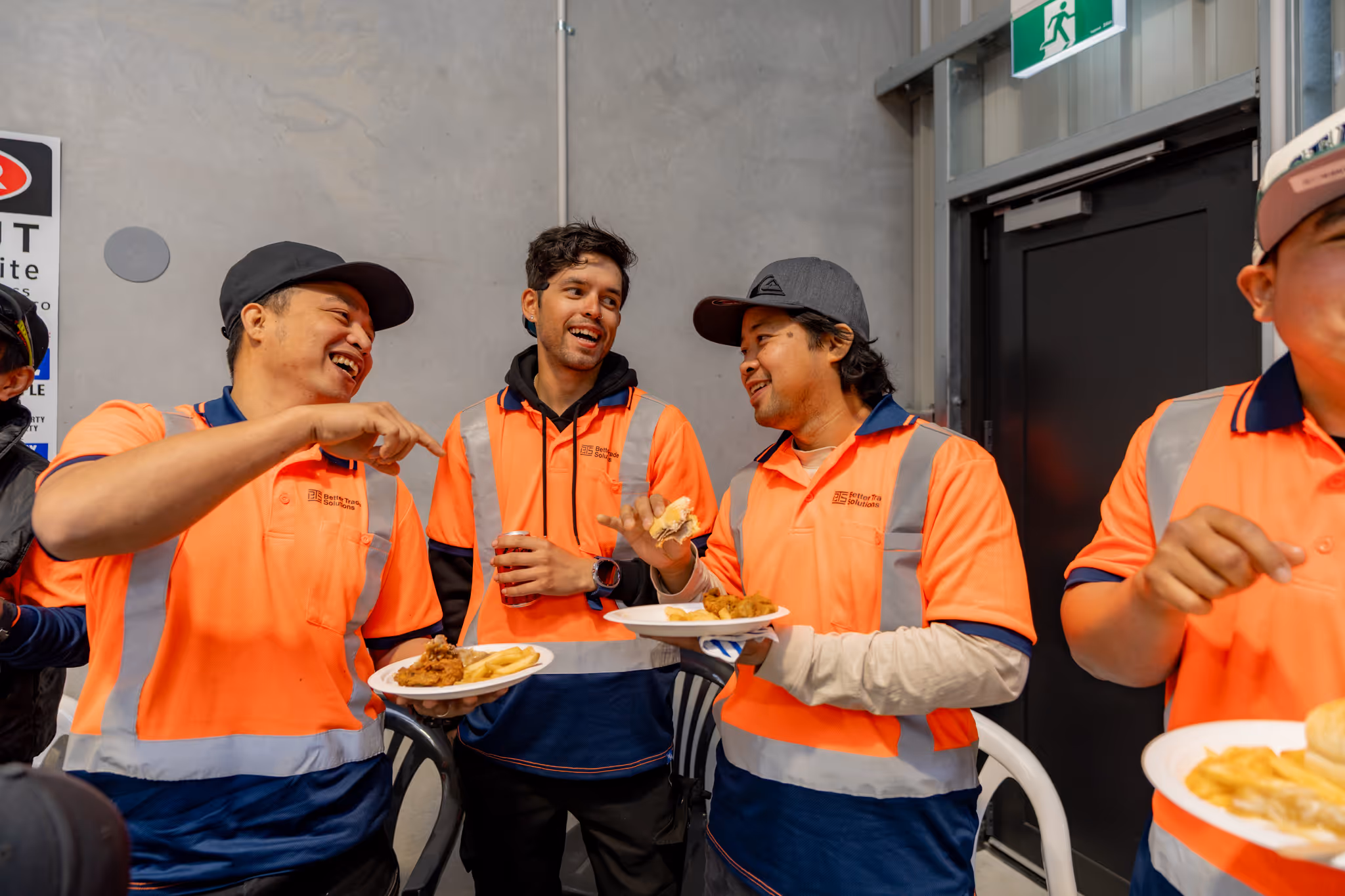Workers in orange high-visibility shirts eating and laughing together in a break room.