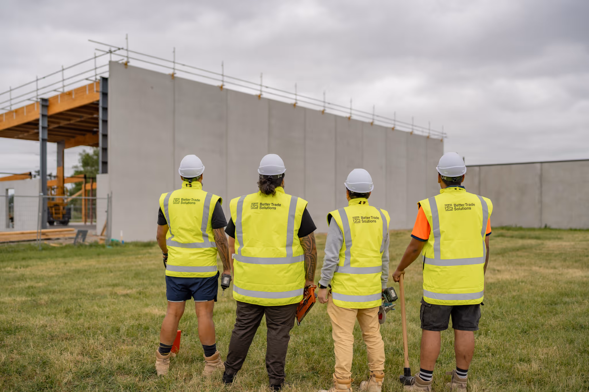 Four construction workers wearing white helmets and yellow Better Trade Solutions vests stand on grass facing a building under construction.