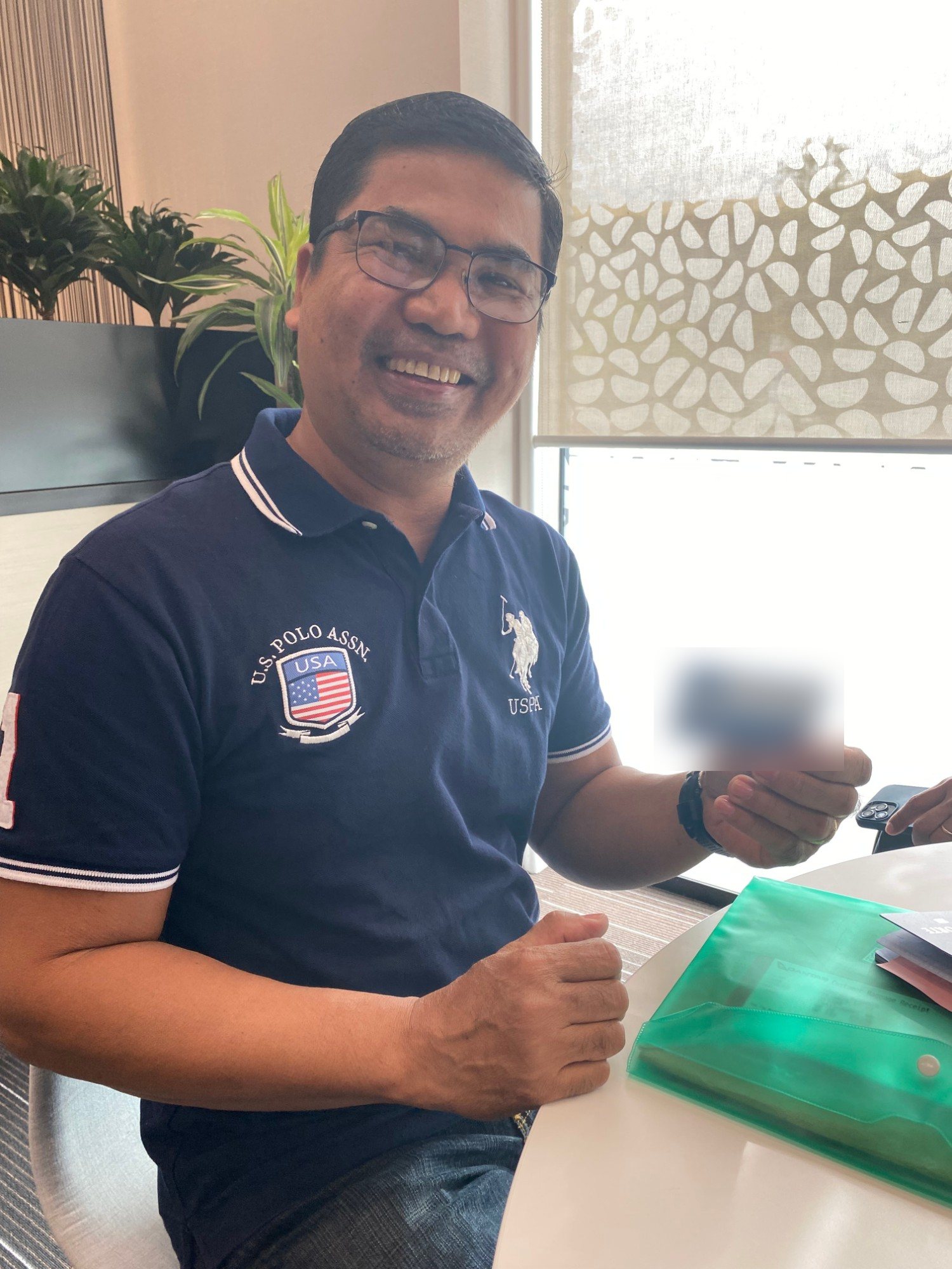 Smiling man in a navy polo shirt sitting at a table and holding up his new New Zealand bank card.