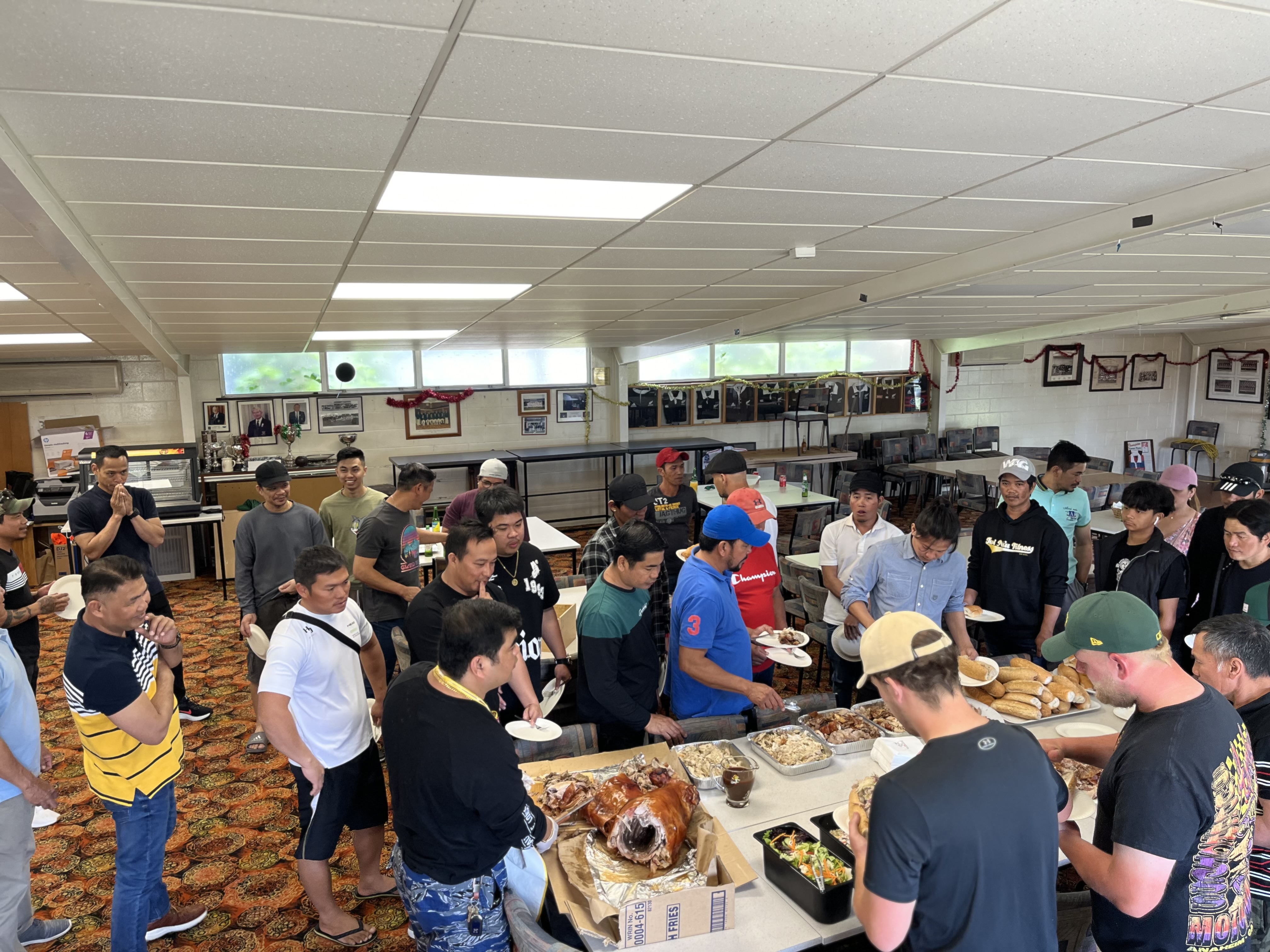 Large group of workers gathered in a sunny backyard for a team barbecue, standing and sitting along a wooden fence.