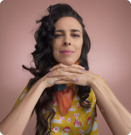 Woman with long wavy dark hair resting her chin on her clasped hands, wearing a yellow shirt with colorful patterns and a multicolored scarf, against a pink background.