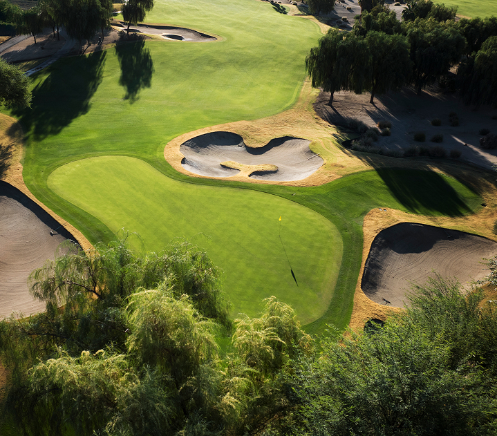 Aerial view of a golf green surrounded by sand bunkers and trees casting shadows.