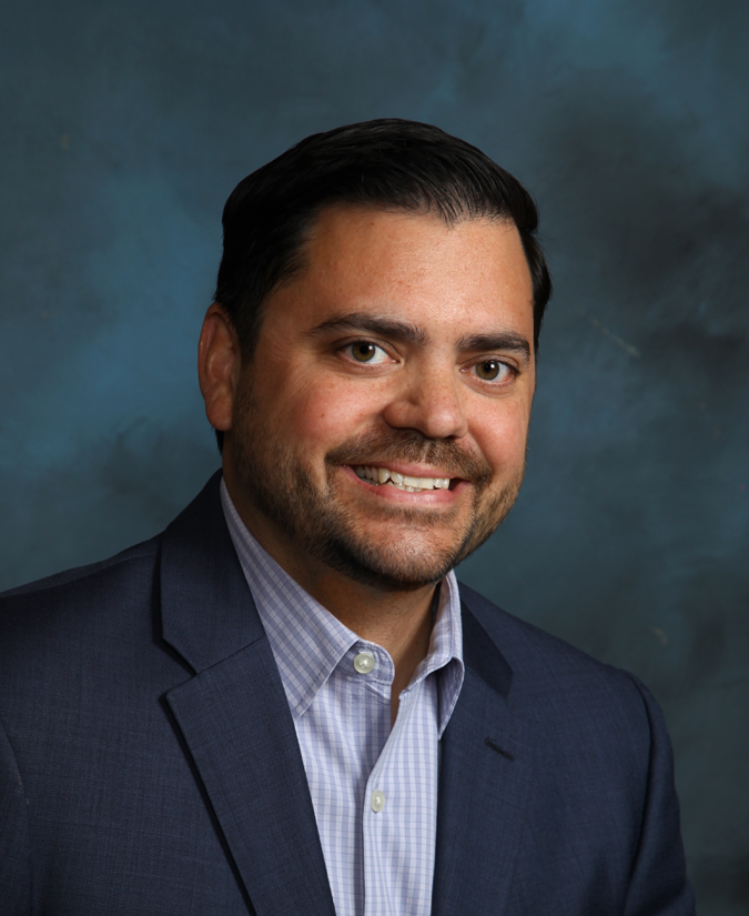 Smiling man with dark hair and beard wearing a blue suit jacket and light checkered shirt against a blue-gray background.
