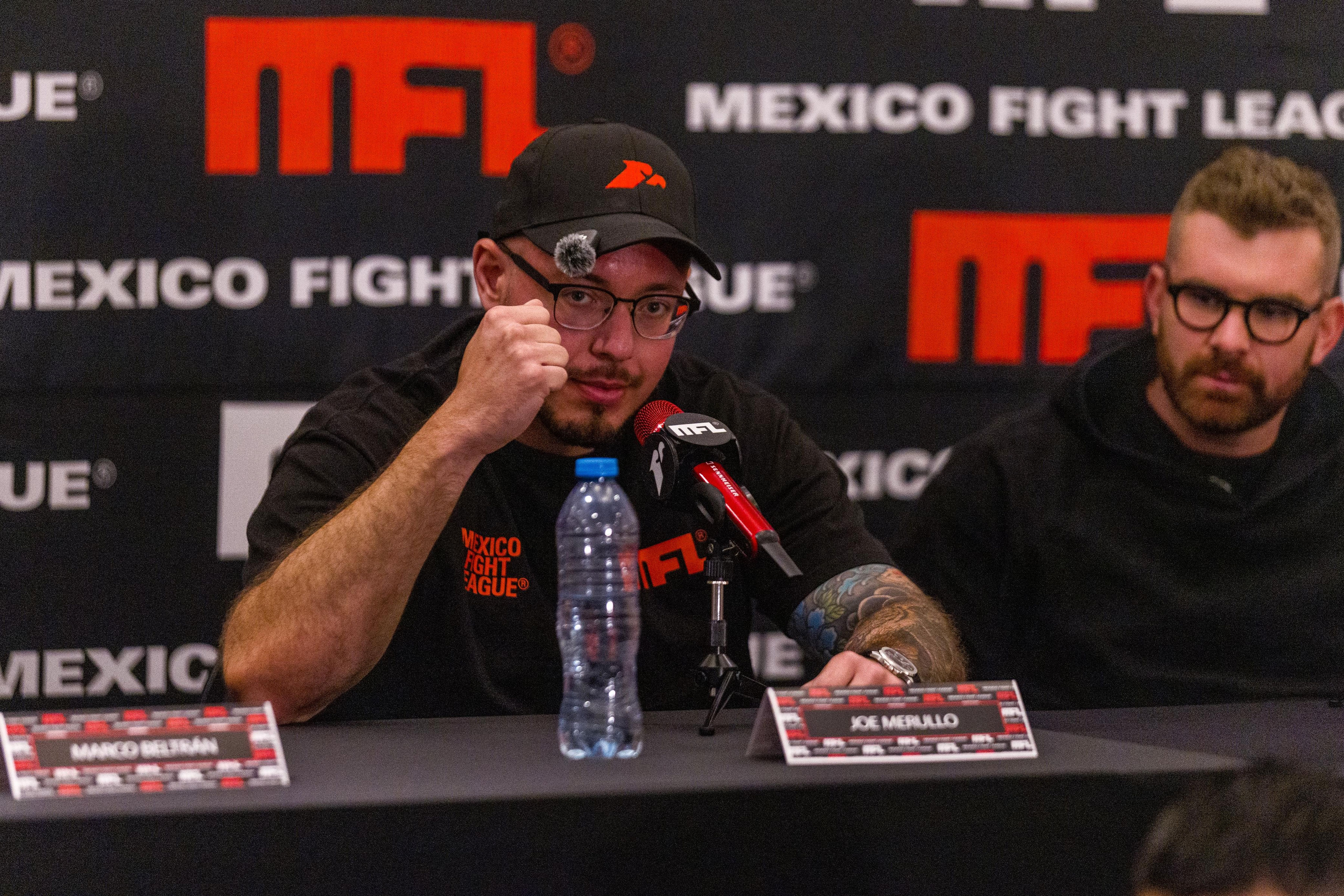 Hombre con gorra y tatuajes levanta el puño frente a micrófono en conferencia de México Fight League.