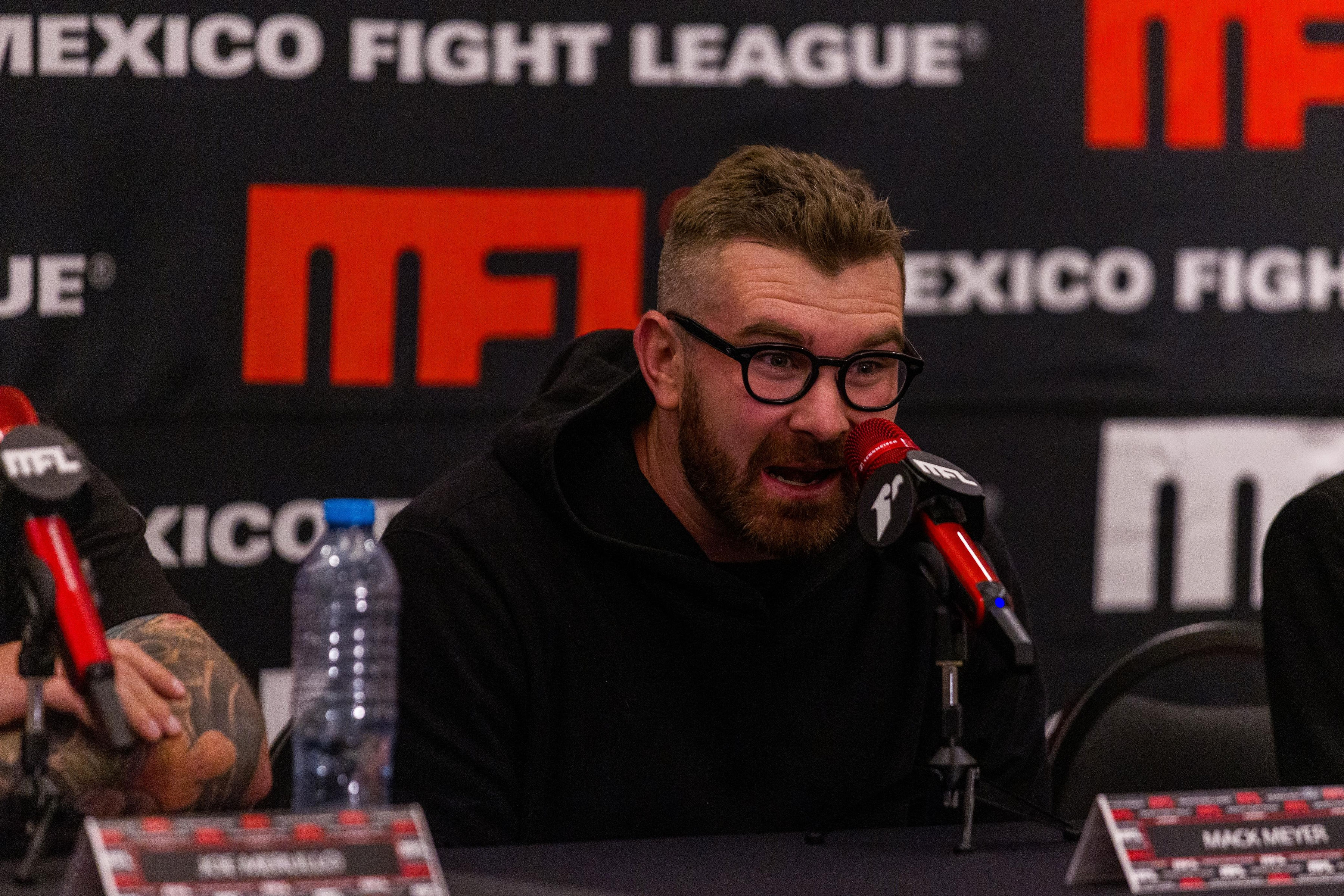 Hombre con gafas y barba hablando en micrófono rojo en conferencia de prensa de Mexico Fight League.