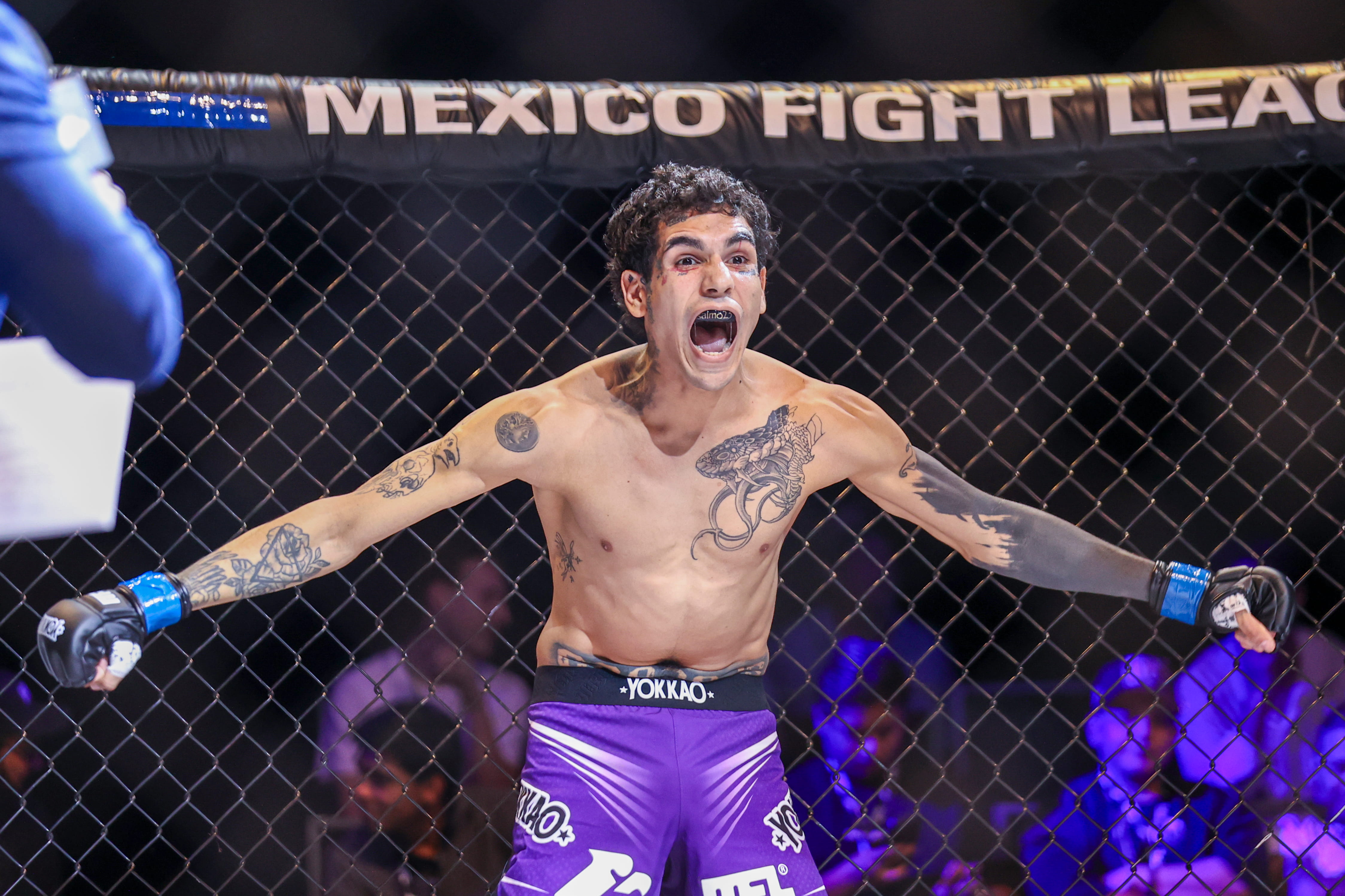 Peleador de artes marciales mixtas con tatuajes y shorts morados celebra dentro del octágono.