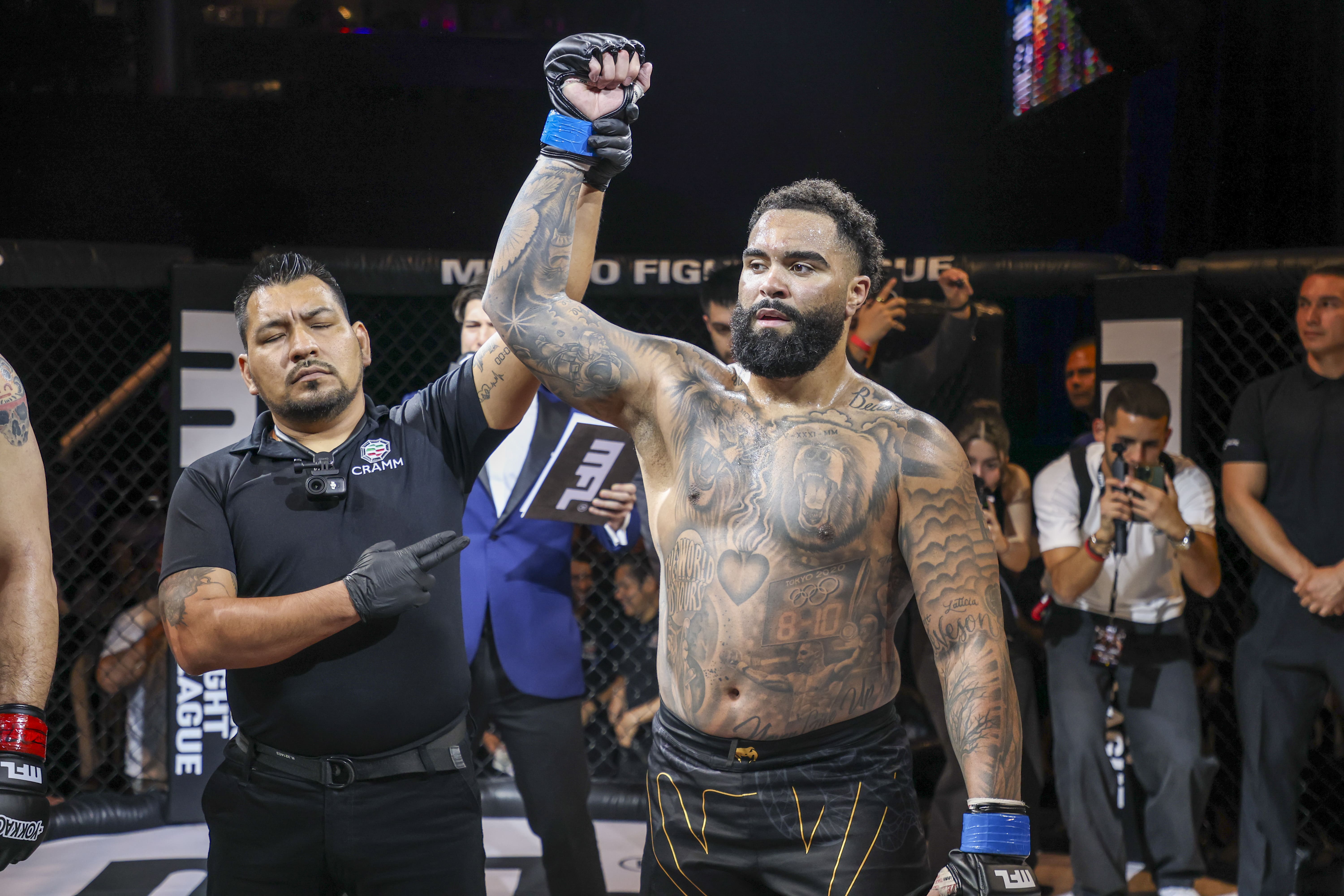 Árbitro levanta la mano de peleador tatuado tras victoria en jaula de pelea profesional.