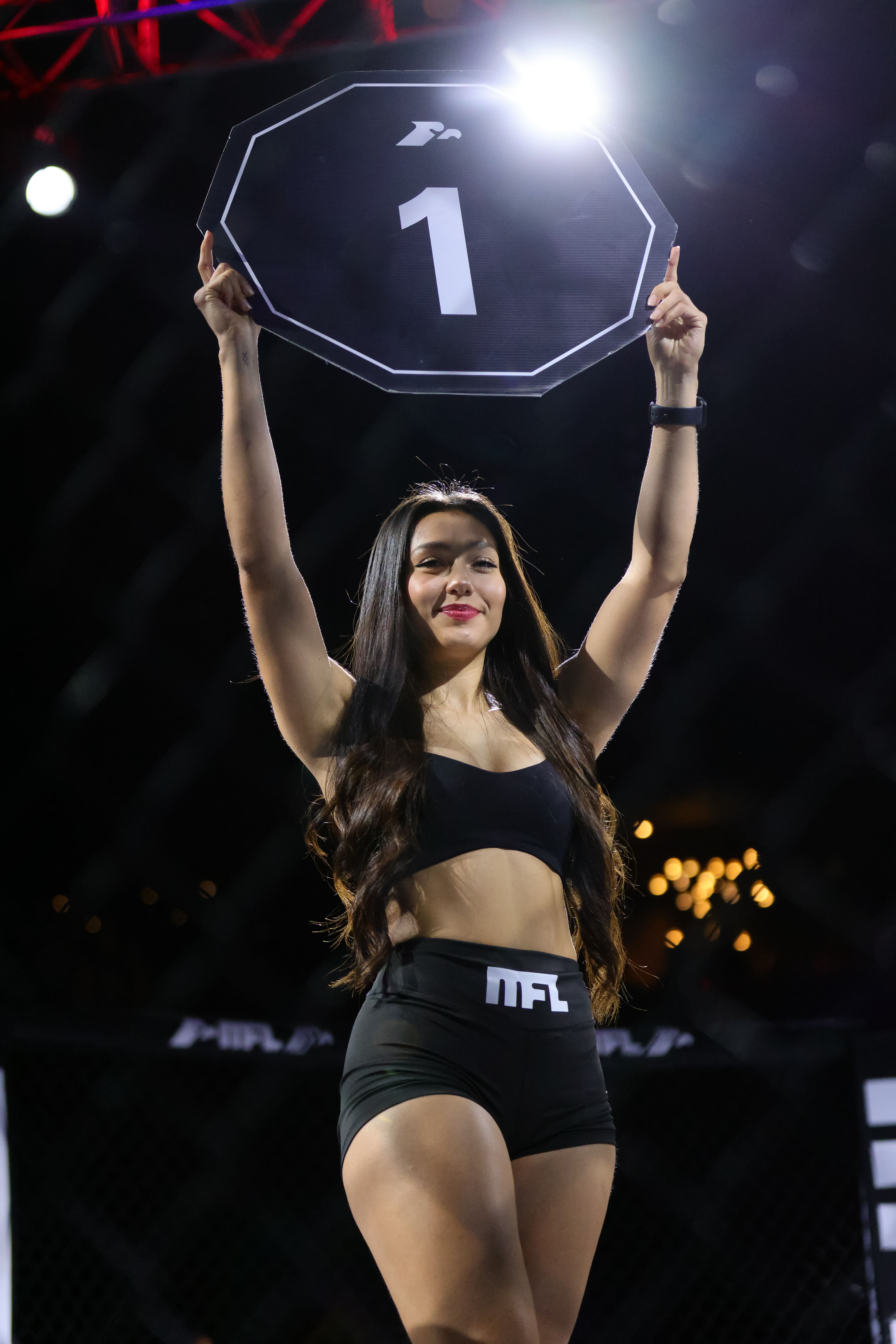 Mujer sosteniendo cartel negro con número uno en un ring de pelea nocturno.