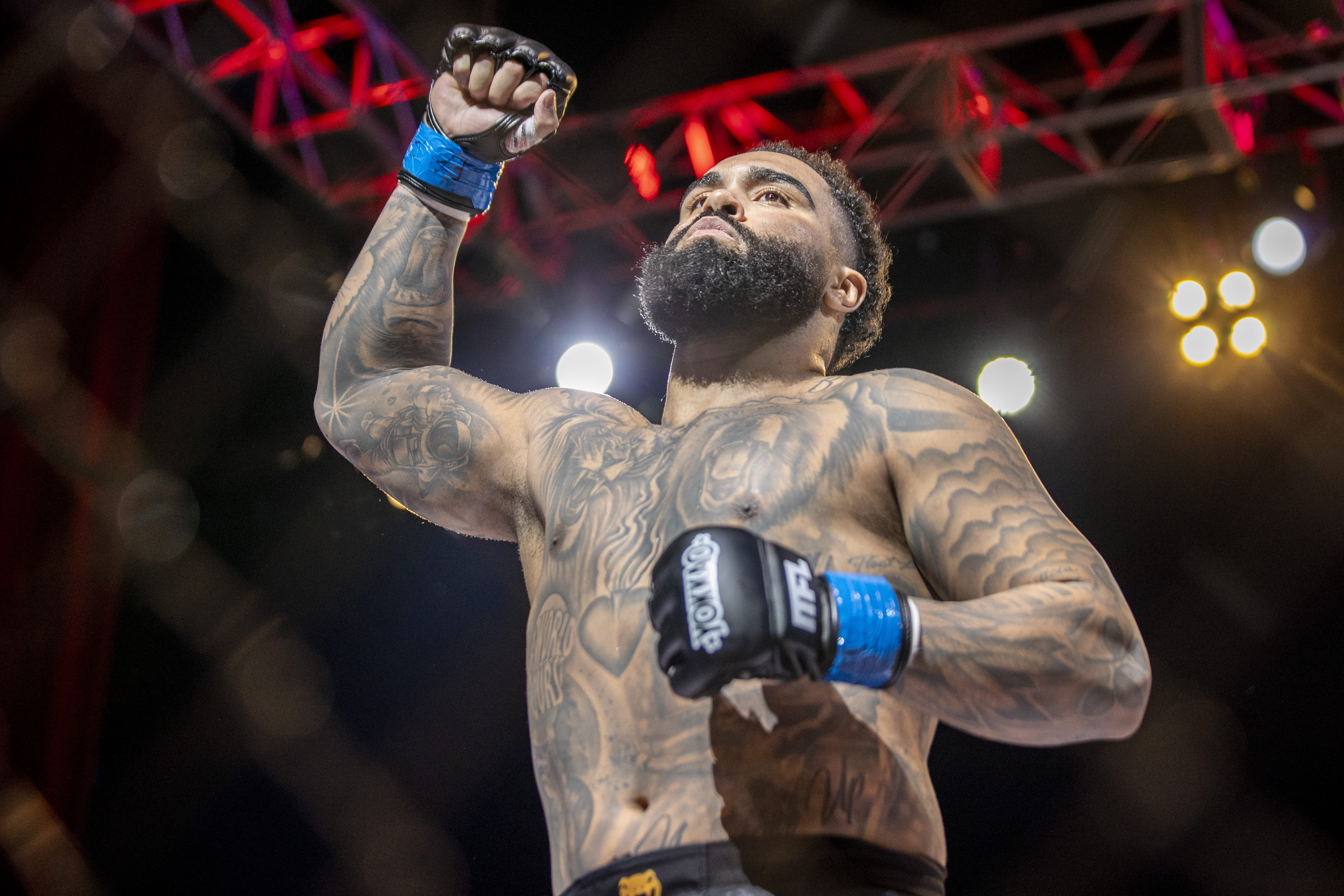 Luchador tatuado y con barba levanta el puño en un ring iluminado con luces rojas y blancas.