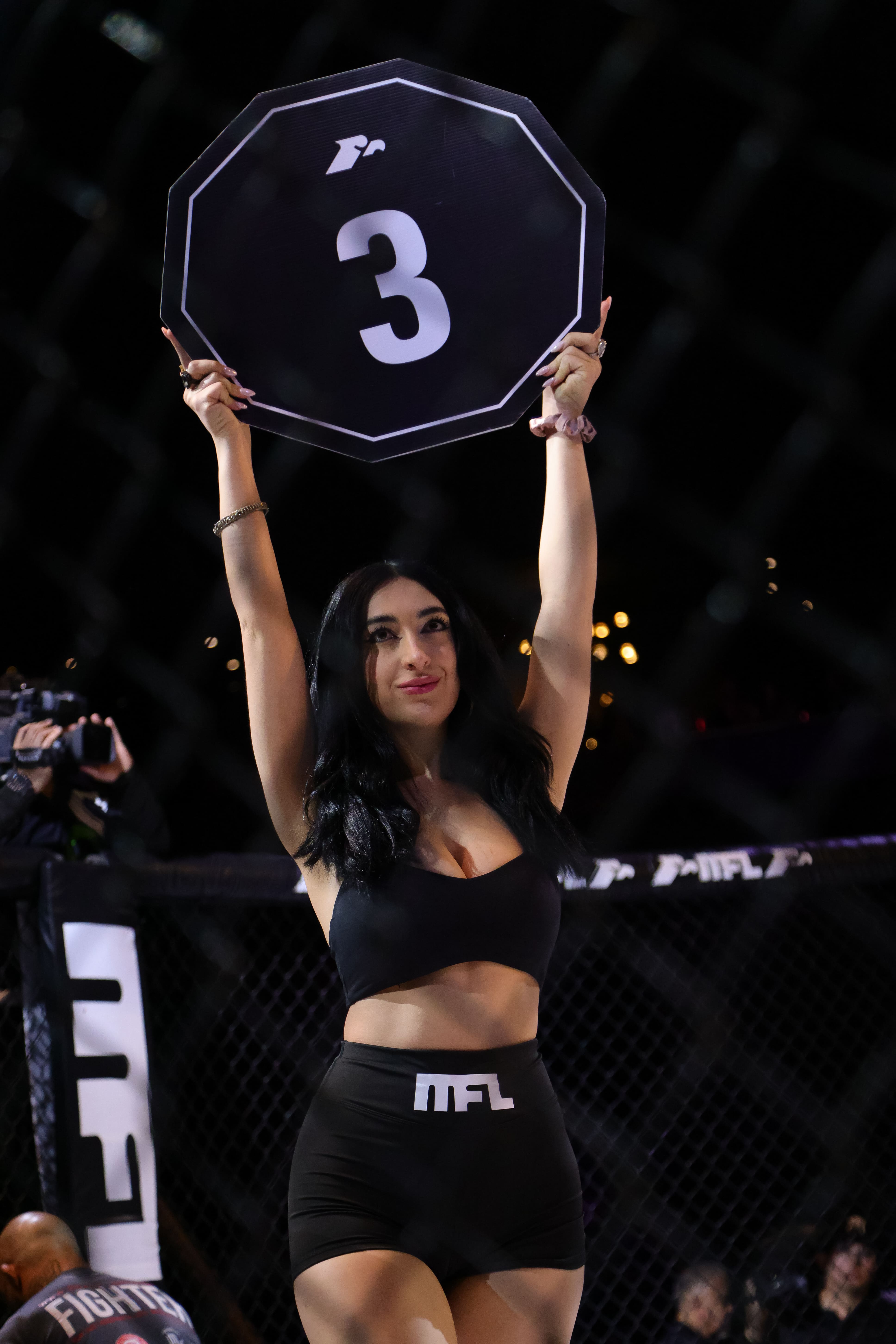 Mujer con ropa negra sostiene cartel octagonal con número 3 en evento deportivo nocturno.