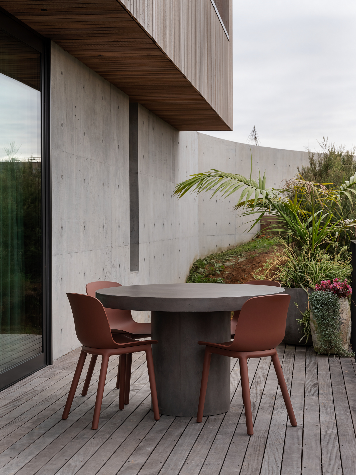 Cantilever House by Matt Deeb Architecture: Modern outdoor patio with a round concrete table and four brown chairs on wooden decking, beside a concrete wall and potted plants.