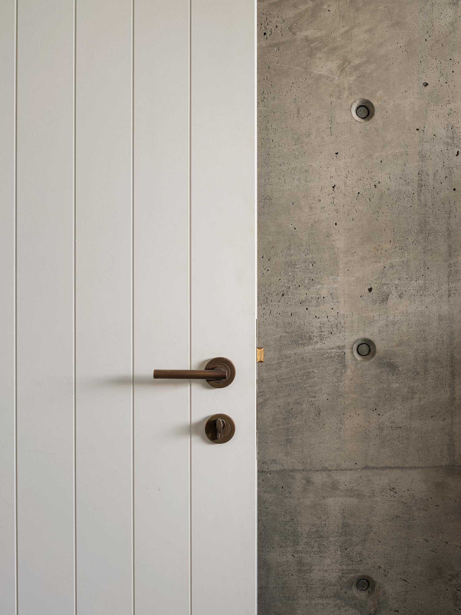 Cantilever House  by Matt Deeb Architecture: Close-up of a white paneled door with a bronze door handle and lock against a textured concrete wall.