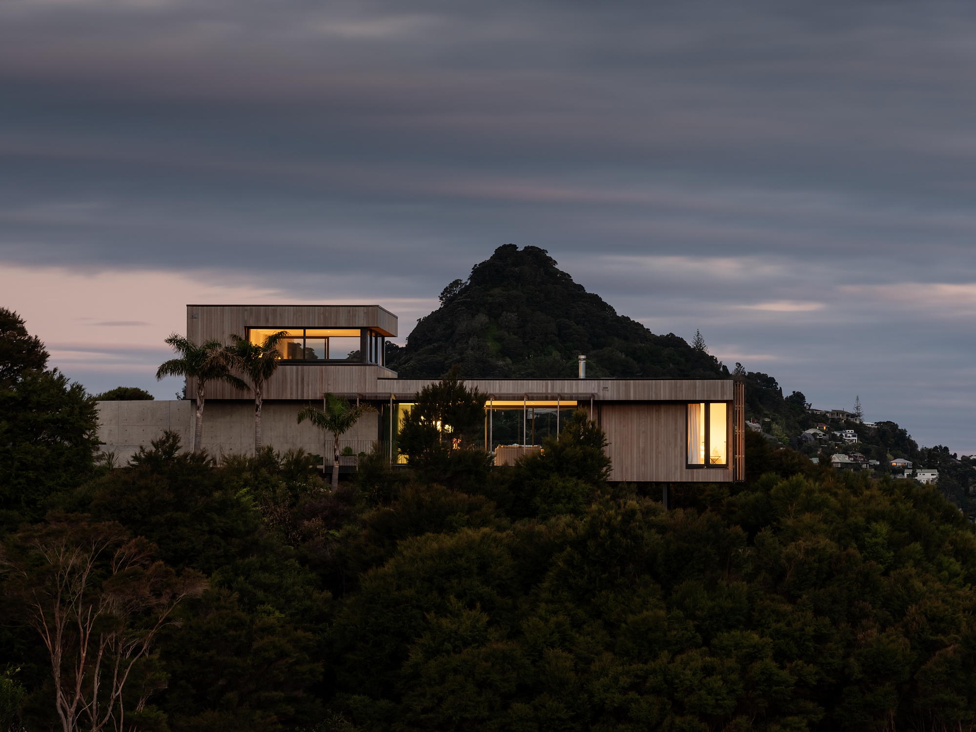 Cantilever House by Matt Deeb Architecture:  Modern house with large windows lit from inside, perched on a wooded hill with a mountain in the background at dusk.