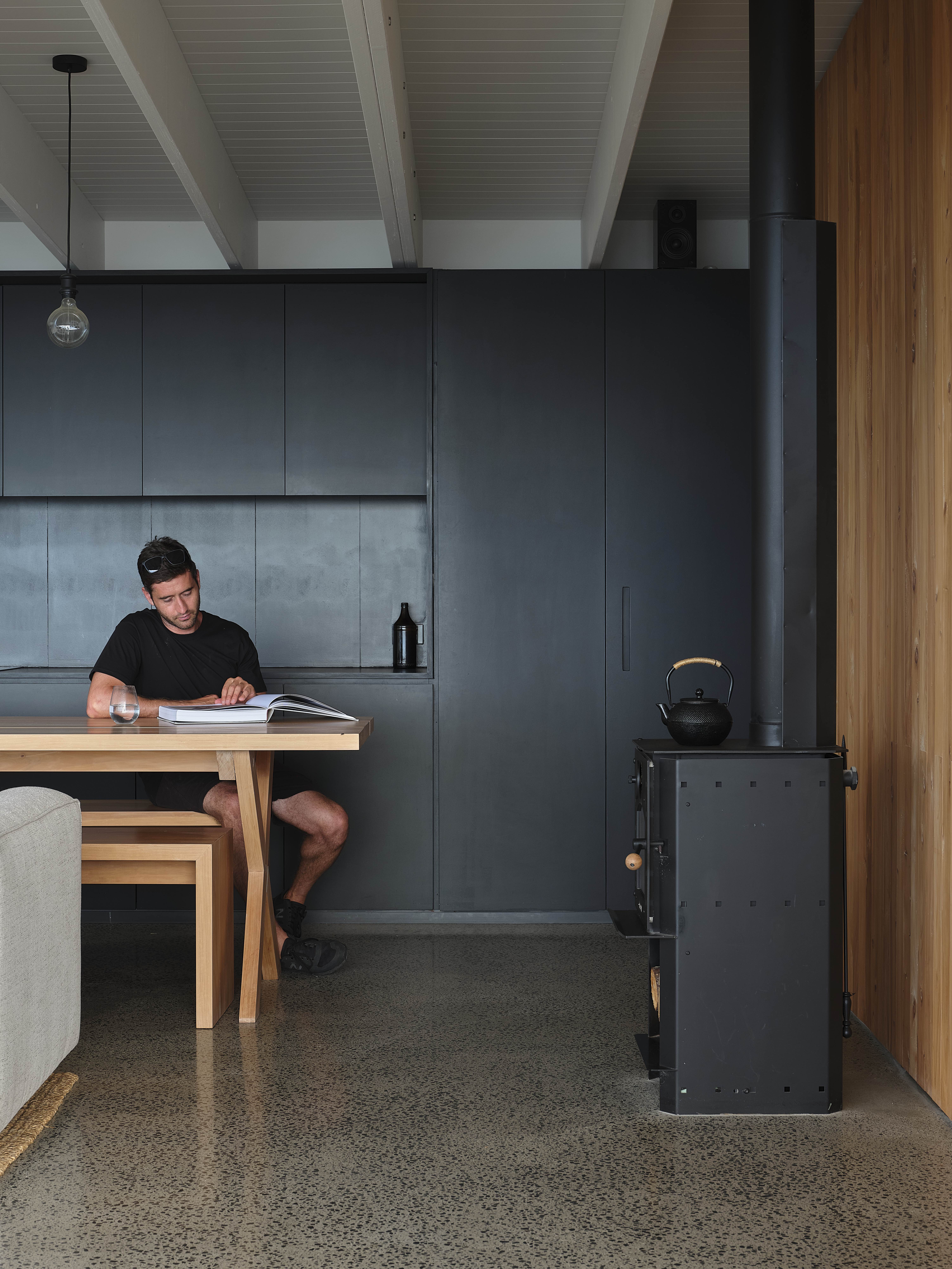 MDA House by Matt Deeb Architecture: Man sitting at a wooden dining table reading a book in a modern kitchen with dark cabinetry and a black wood stove.