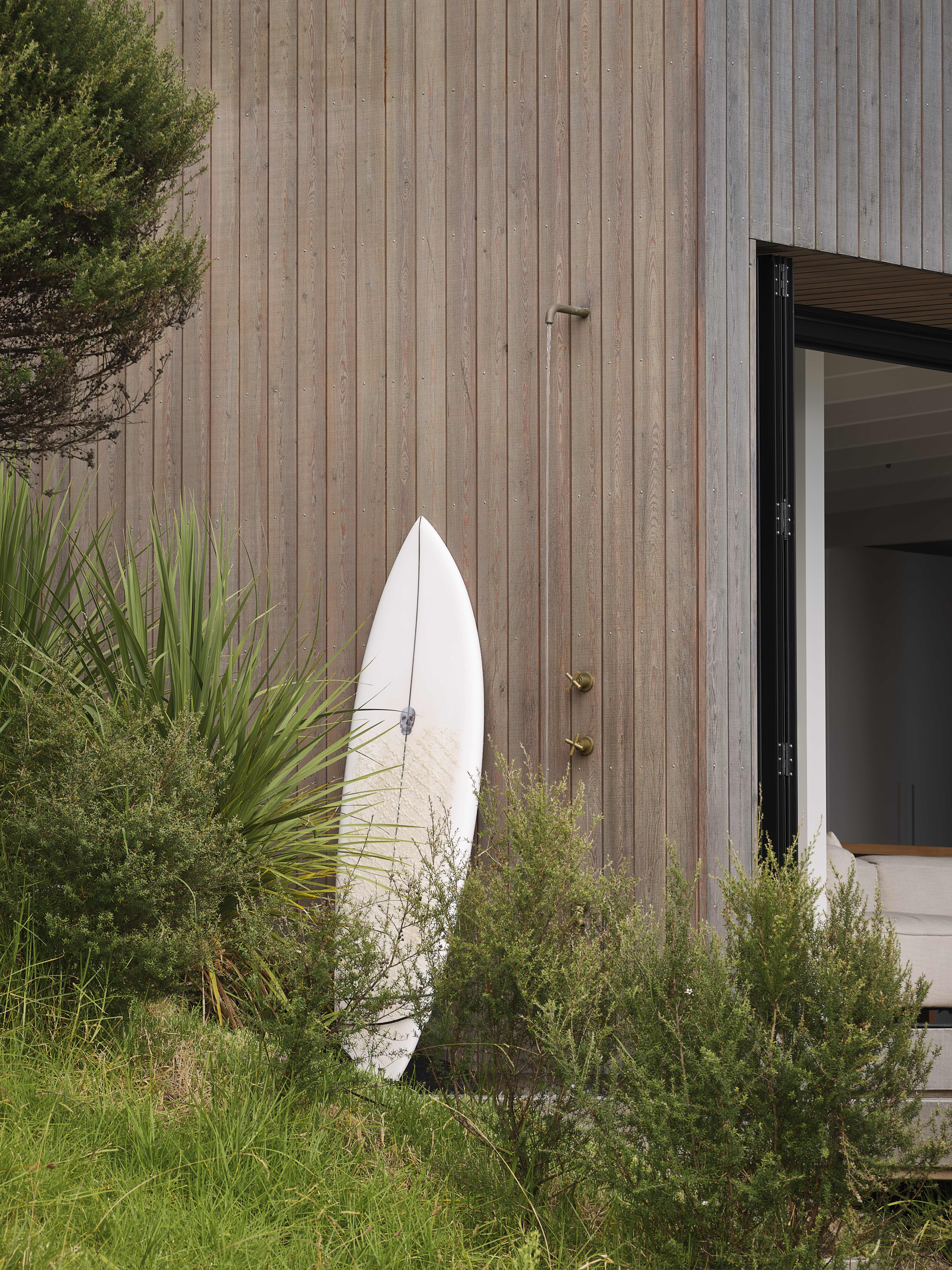 MDA House by Matt Deeb Architecture: White surfboard leaning against a wooden wall next to outdoor shower, with green bushes and grass in front and a partially open door showing a beige couch inside.