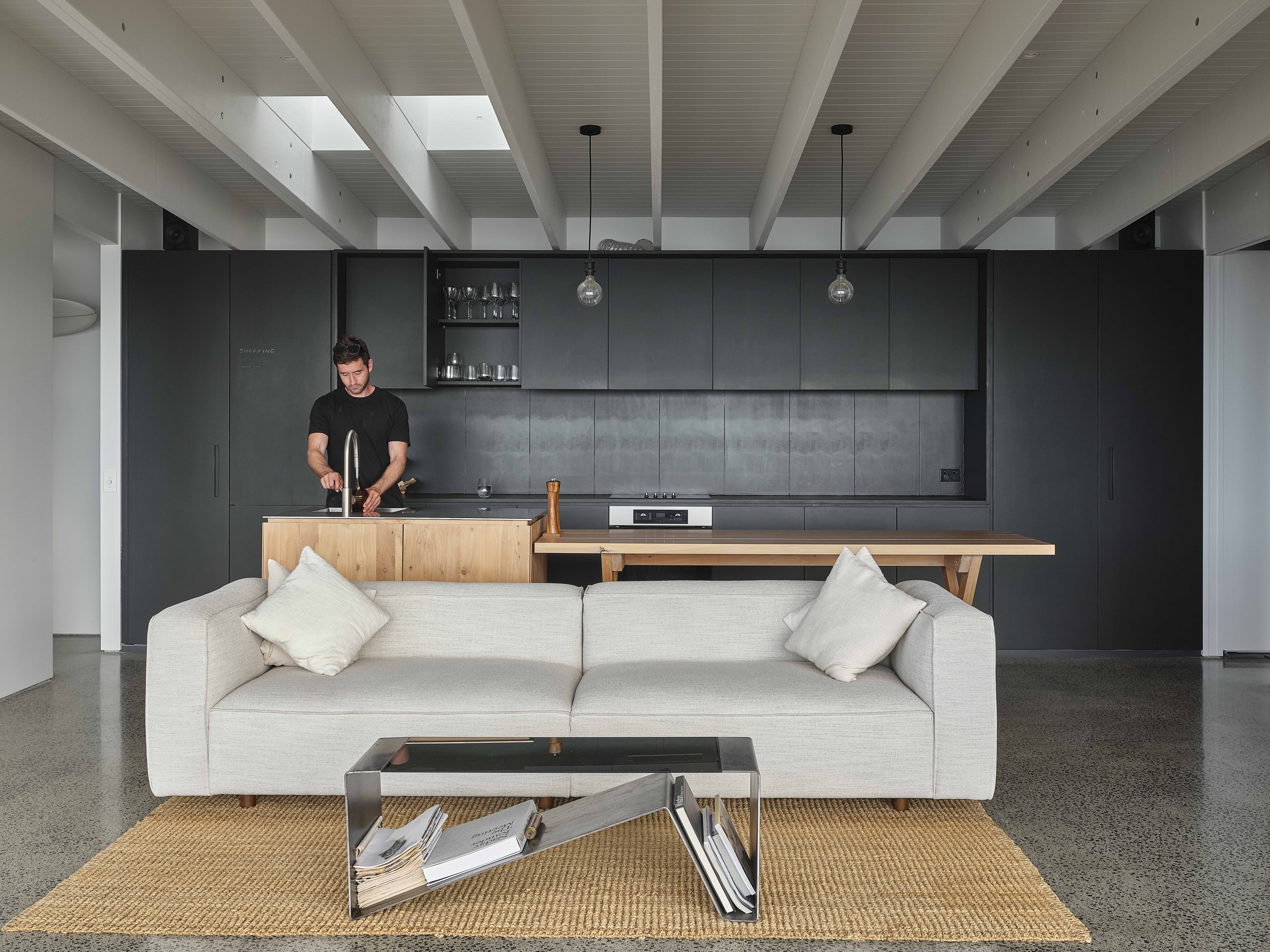 MDA House by Matt Deeb Architecture: Modern open living space with a light gray sofa and a glass coffee table on a woven rug, and a man washing hands in a kitchen with black cabinets and wooden island in the background.