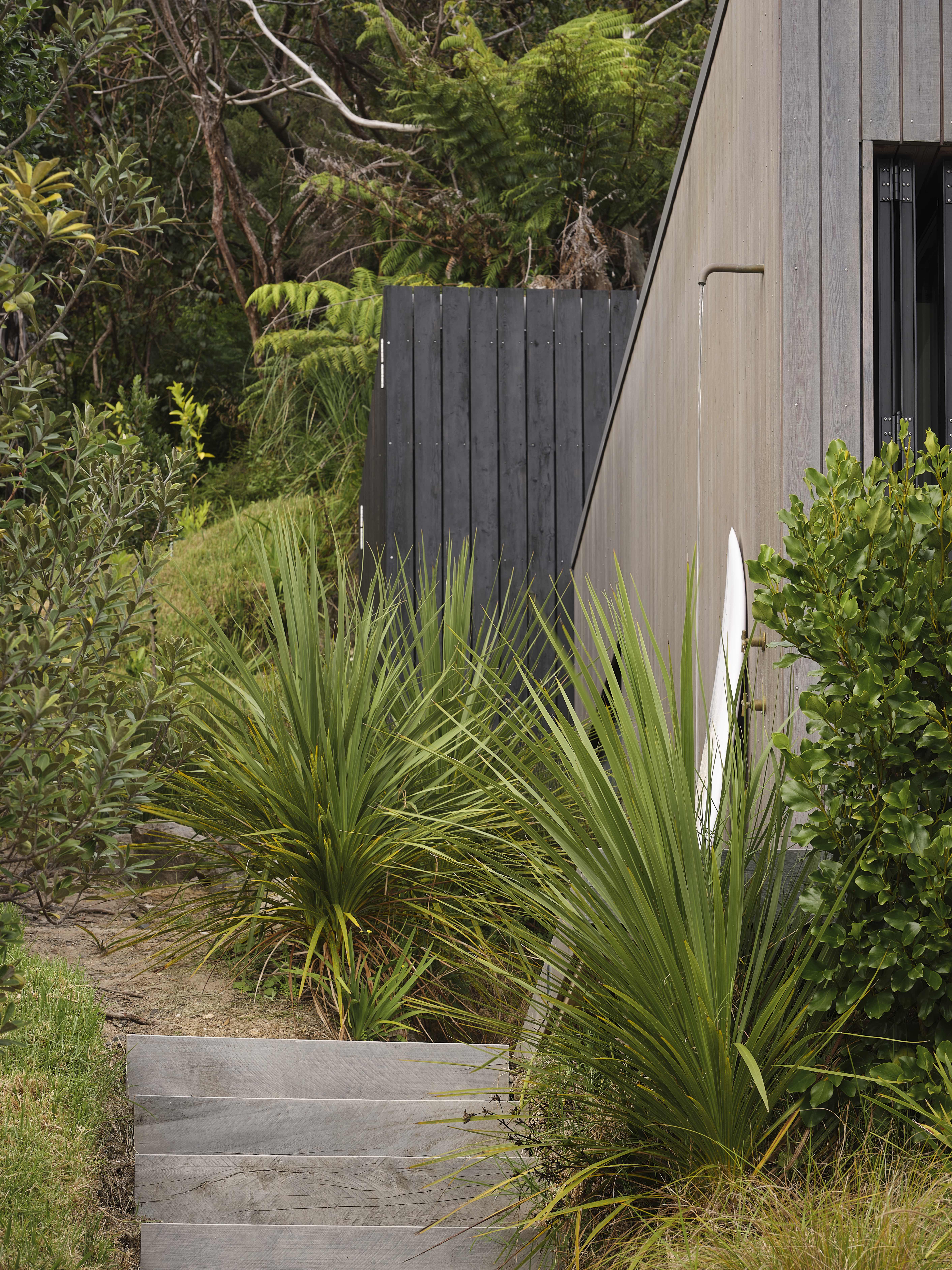 MDA House by Matt Deeb Architecture: Outdoor wooden steps leading up to a black wooden shed, surrounded by green plants and next to a modern building with an outdoor shower.
