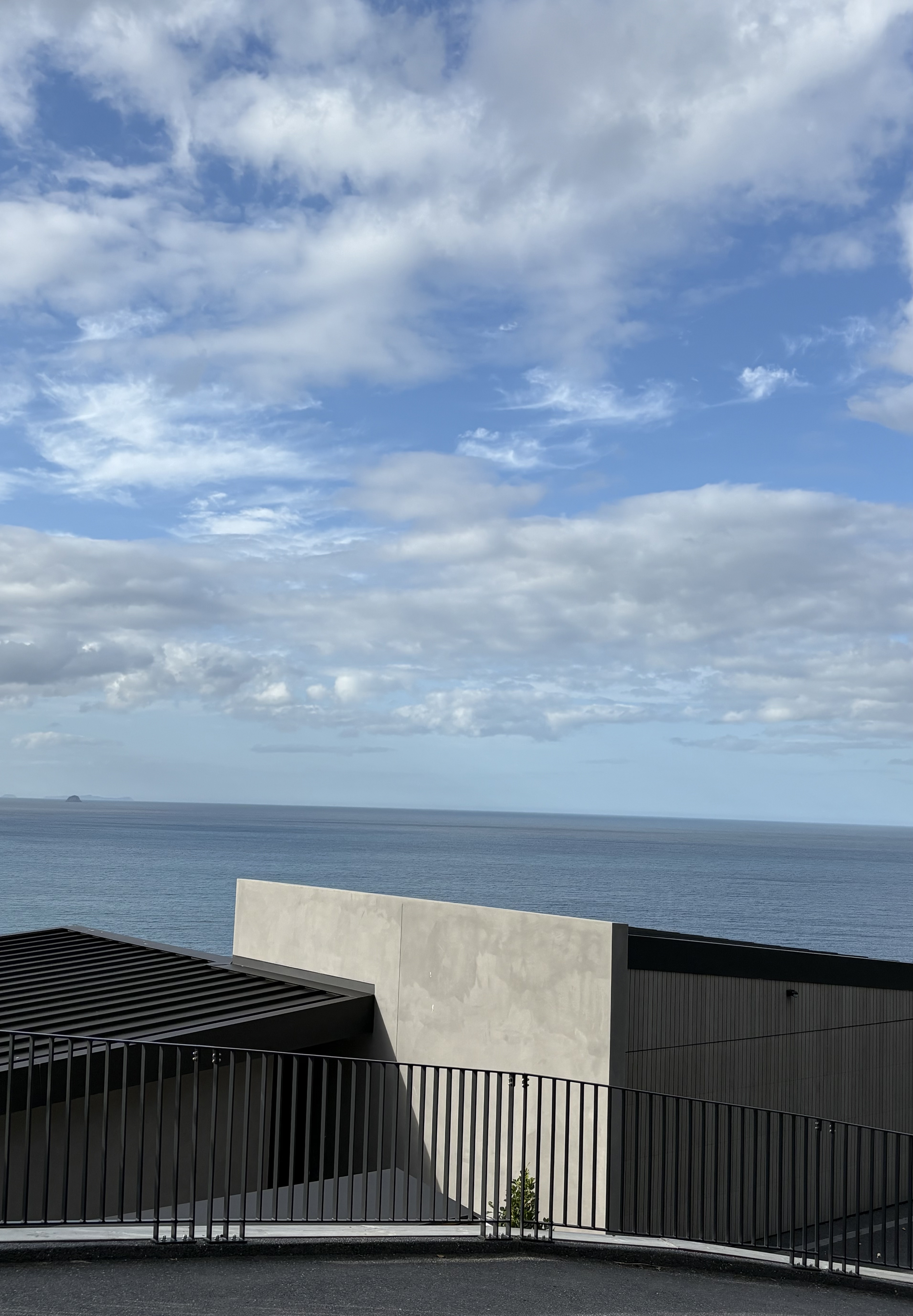 Fantail House by Matt Deeb Architecture: View of a calm ocean under a partly cloudy blue sky with modern building rooftops and a black metal railing in the foreground.