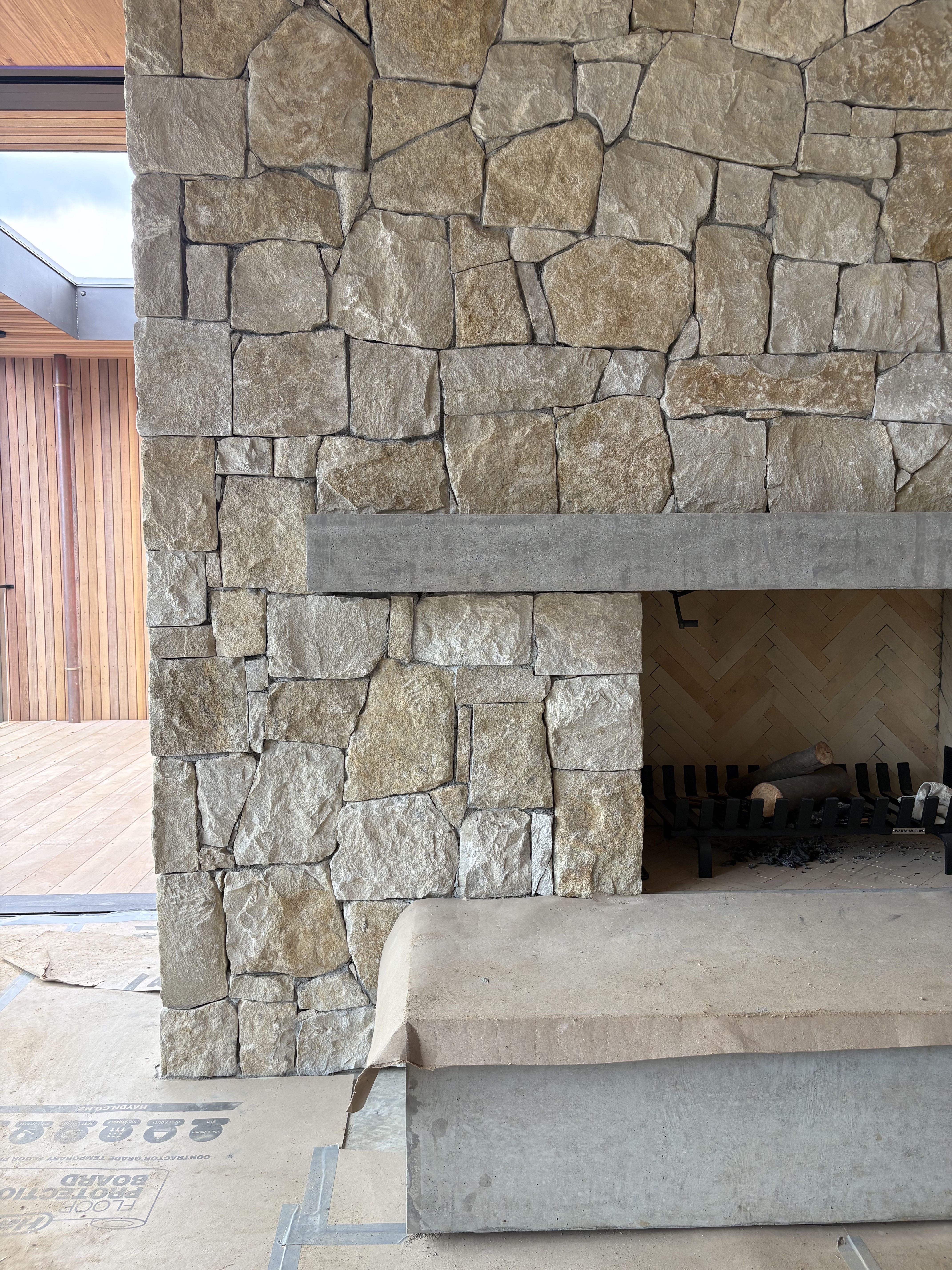 Tirinui house by Matt Deeb Architecture: under construction Stone wall with a concrete fireplace and a fire grate holding logs inside, next to a wooden deck area.