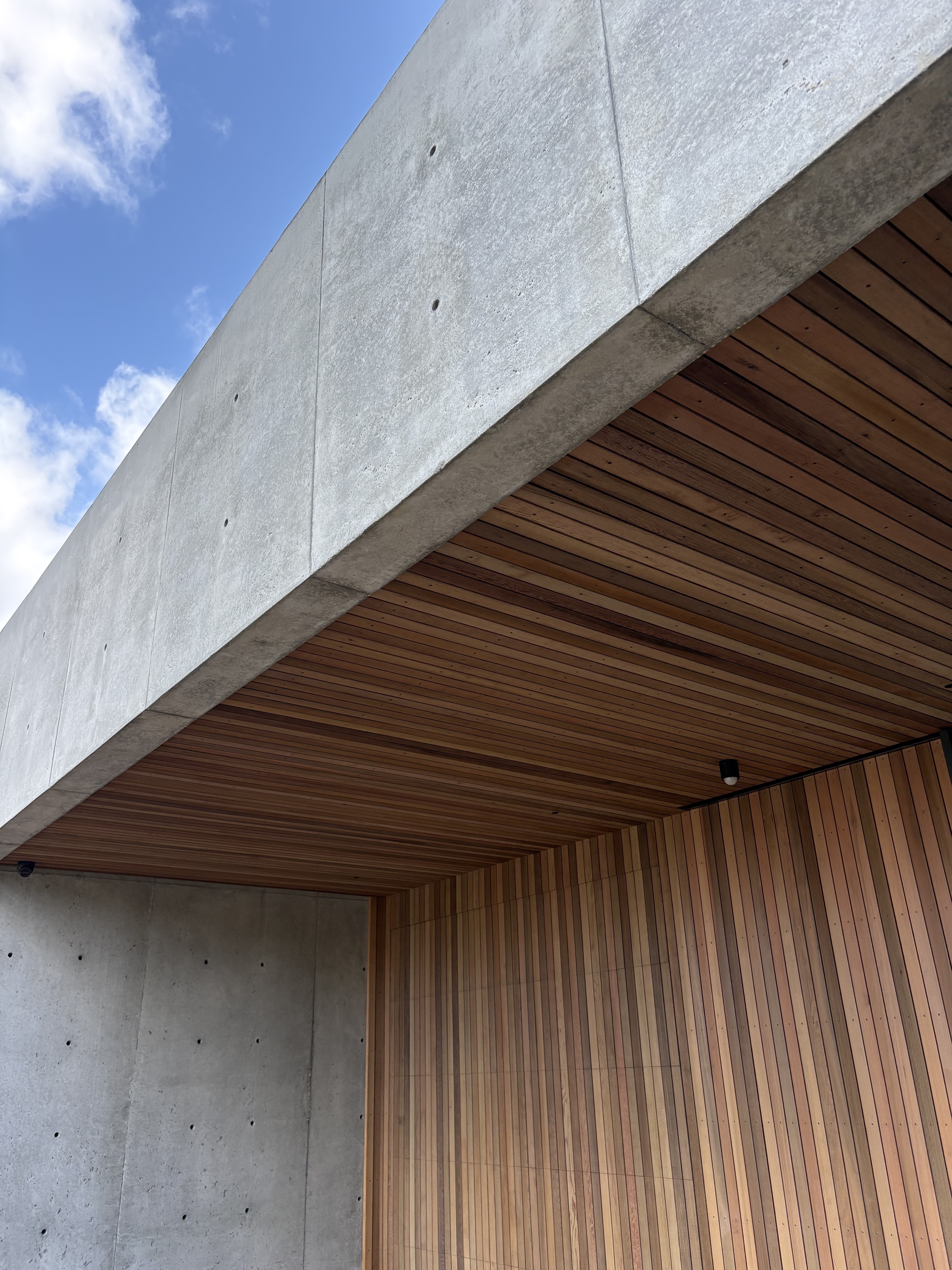Tirinui House by Matt Deeb Architecture: Modern building corner with smooth gray concrete walls and a wooden ceiling and wall under a blue sky with clouds.