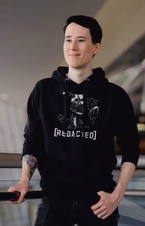 Athan wearing a black hoodie with a graphic design and text '[REDACTED]' standing indoors with one hand on a railing.