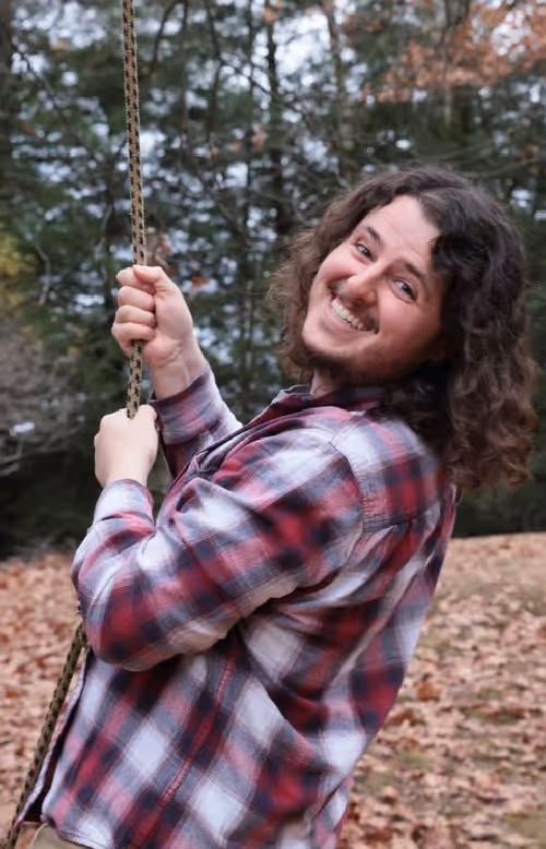 Smiling man with curly hair wearing a red and white plaid shirt holding onto a rope swing outdoors with fallen autumn leaves on the ground.