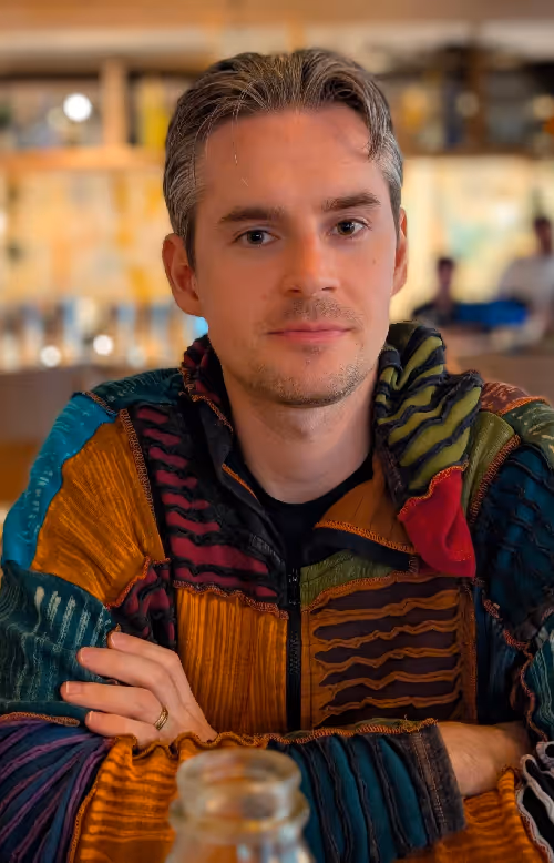 Derek in a multicolored patchwork jacket sitting at a table indoors with arms crossed.