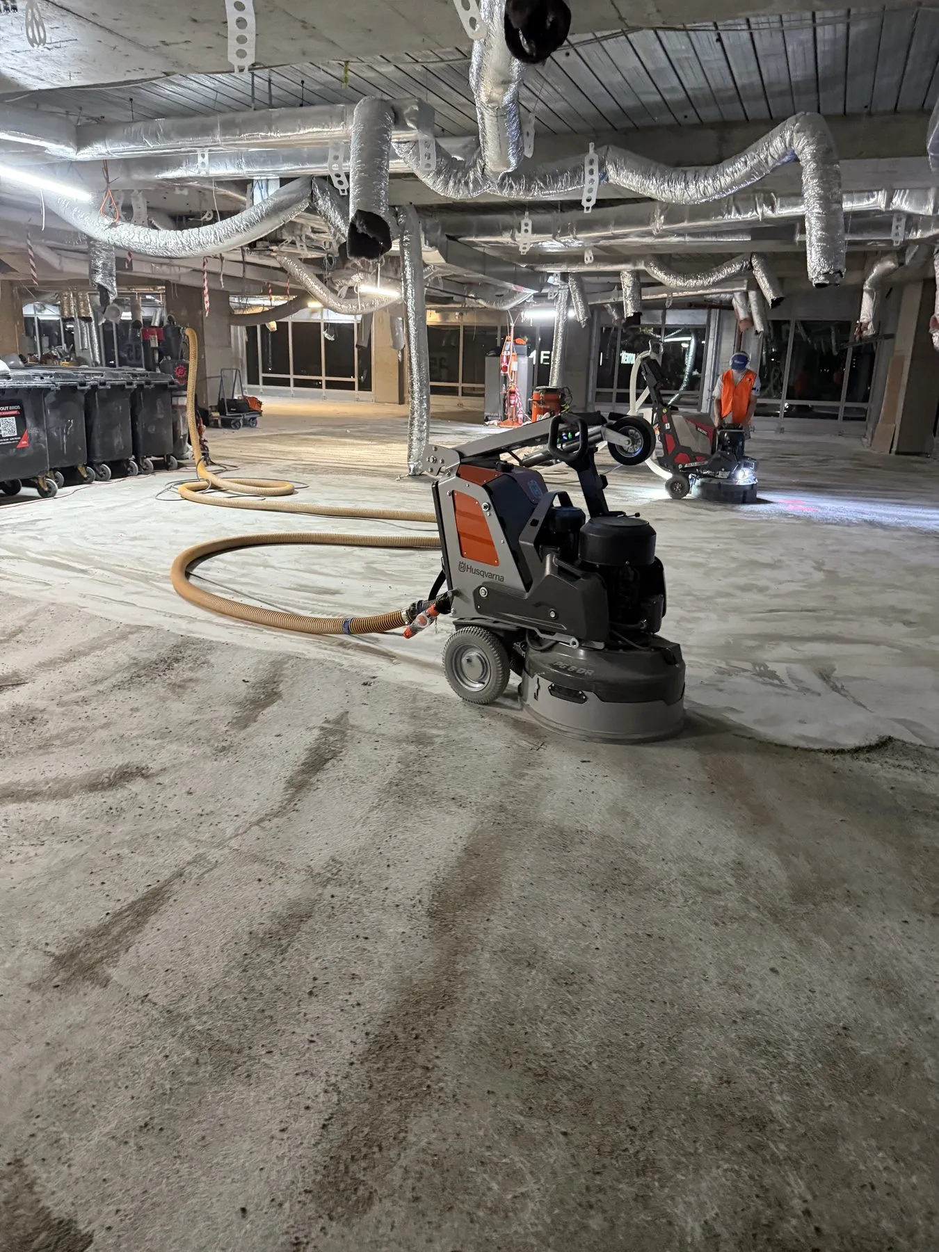 Two floor grinding machines clearing adhesive from a large concrete floor in an industrial building.
