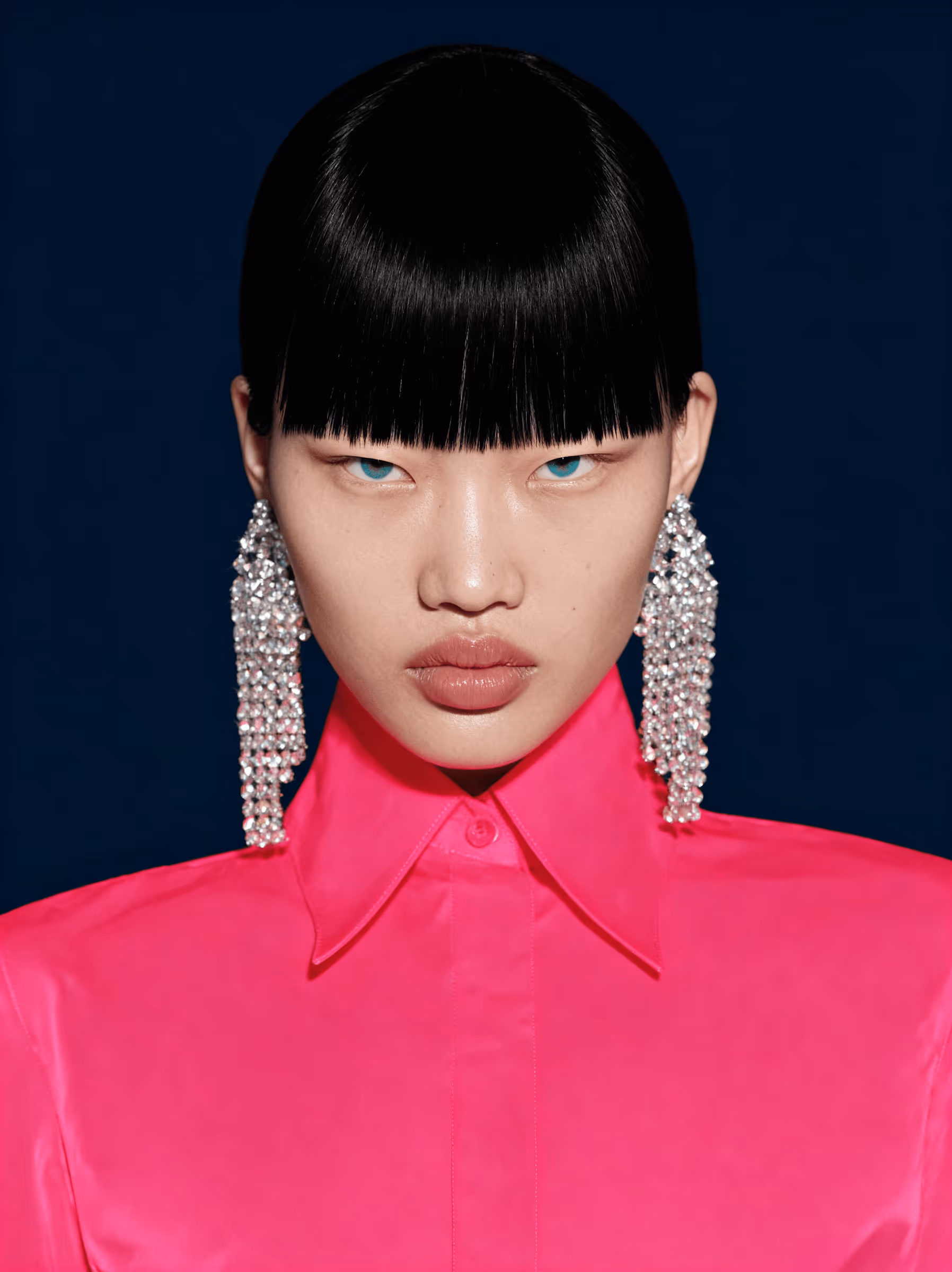 Fashion model with sharp black bangs, blue eyes, wearing large sparkling earrings and a bright pink collared shirt against a dark background.