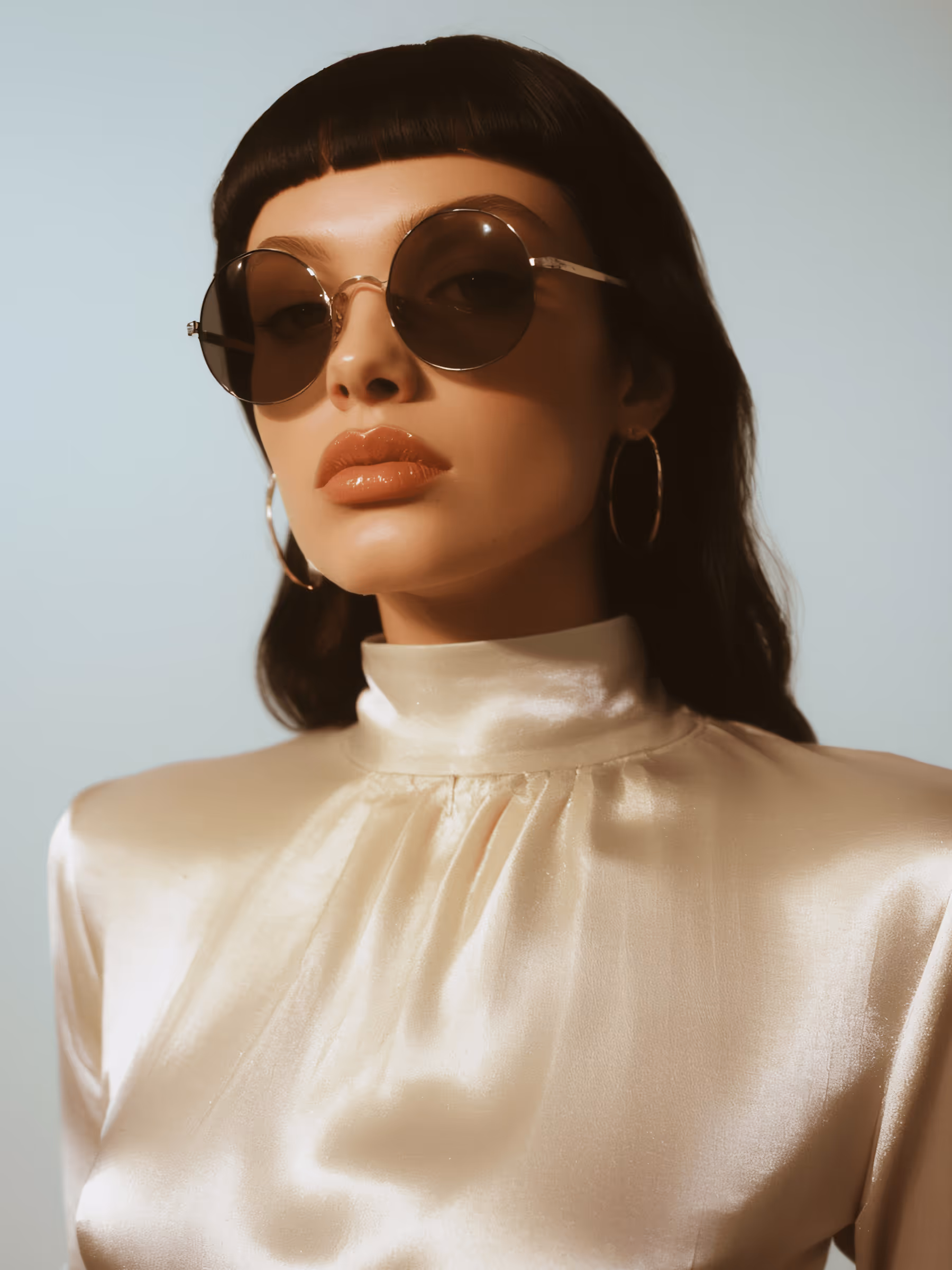 Woman with blunt bangs wearing round sunglasses, gold hoop earrings, and a shiny cream satin high-neck blouse.