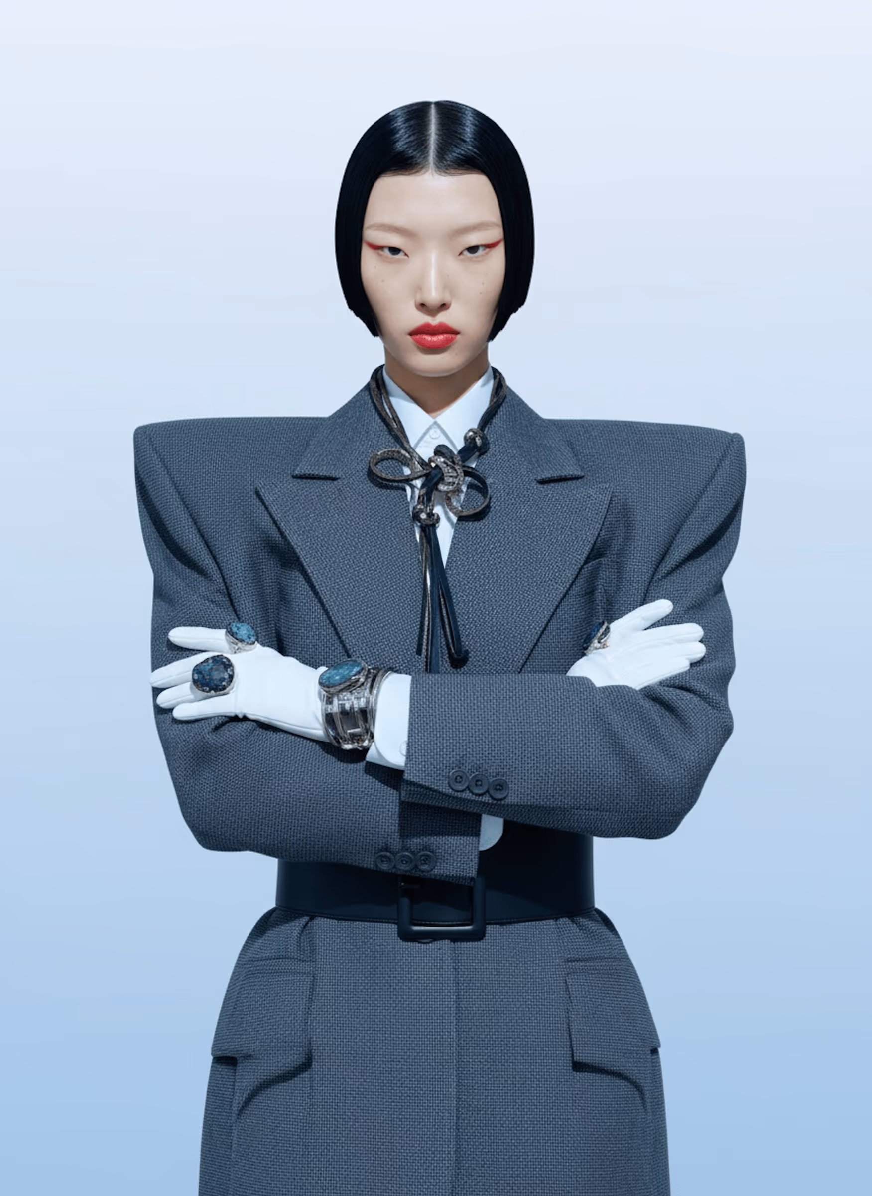 Fashion model with sleek black bob haircut, wearing a gray structured blazer with shoulder pads, white gloves, and large gemstone rings, posing with arms crossed.