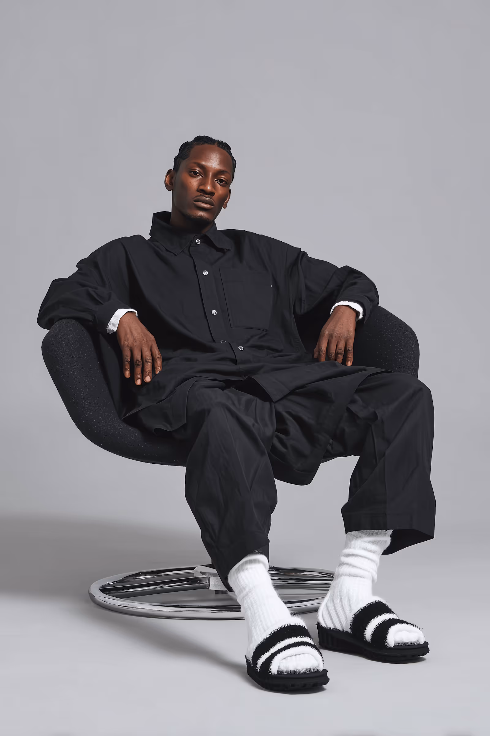 Man with braided hair wearing black oversized shirt and pants, white socks, and black-and-white slides sitting on a modern black chair with a chrome base against a gray background.