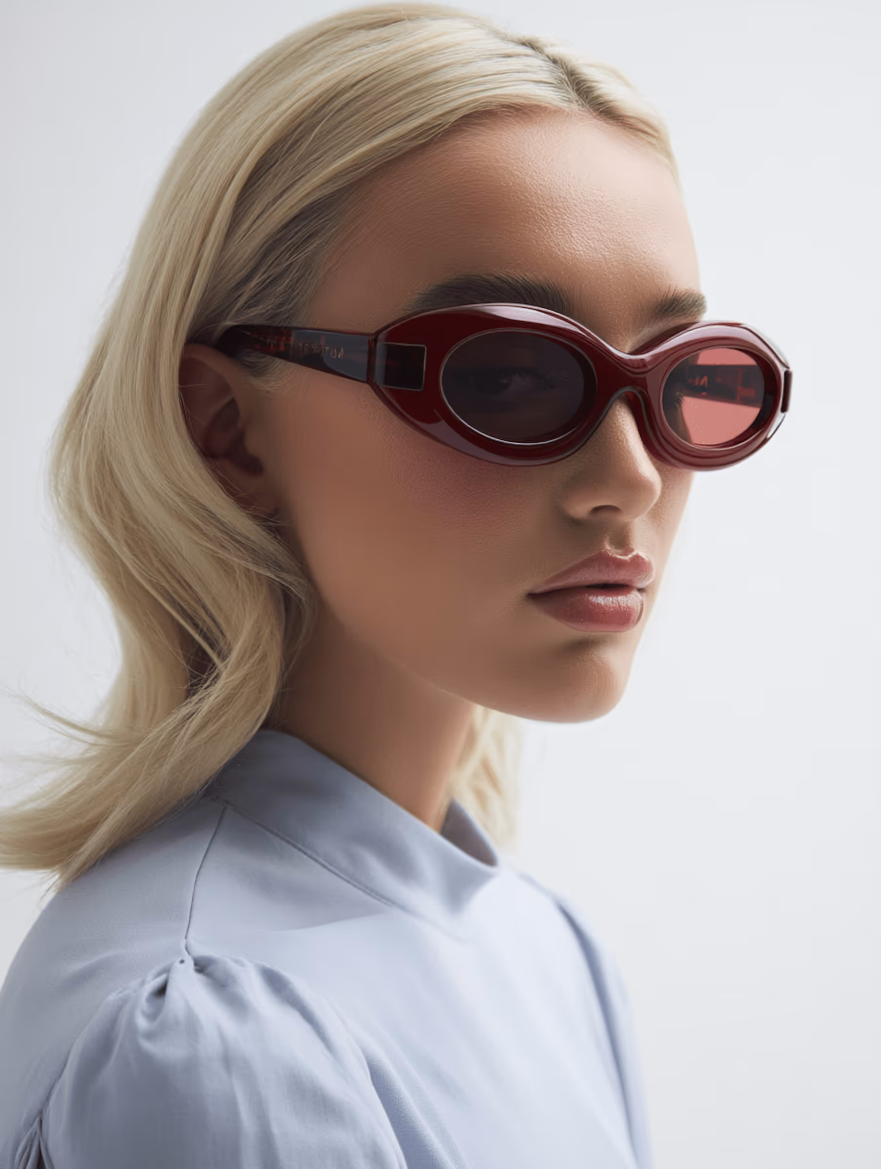 Blonde woman wearing red oval sunglasses and a light blue blouse against a white background.