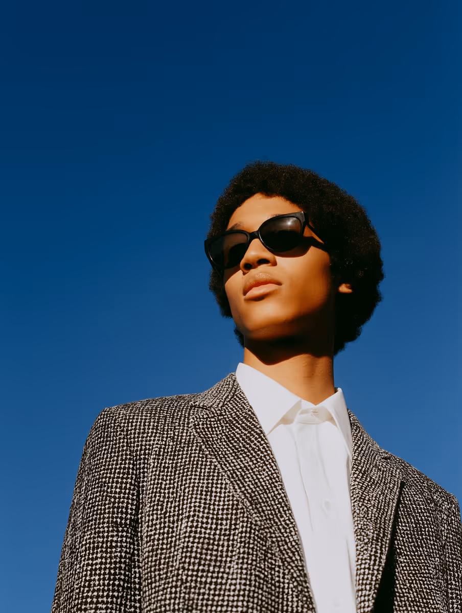 Young man wearing sunglasses and a black-and-white patterned blazer against a clear blue sky.