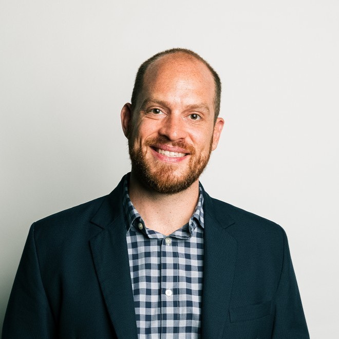 Smiling man with a beard wearing a blue blazer over a checked shirt, standing against a neutral background.