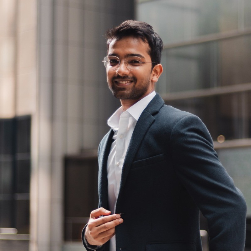 Young man in a suit and glasses, smiling confidently outdoors with modern buildings in the background.