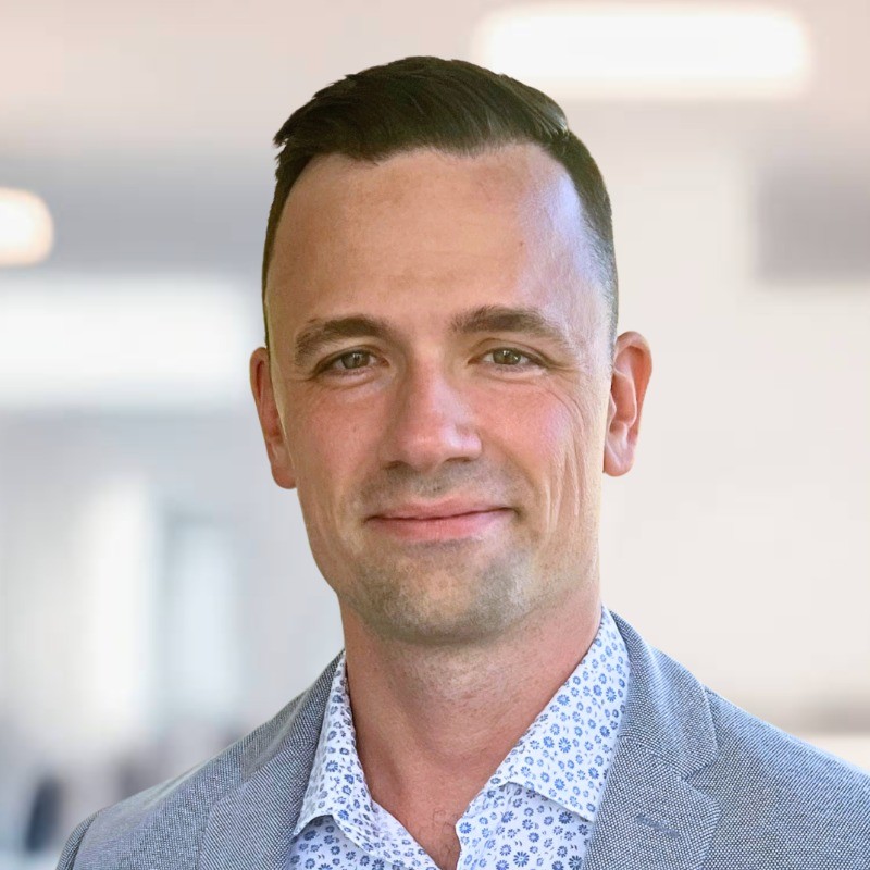 A man with short hair and a subtle smile is wearing a patterned shirt and blazer, against a softly blurred background.